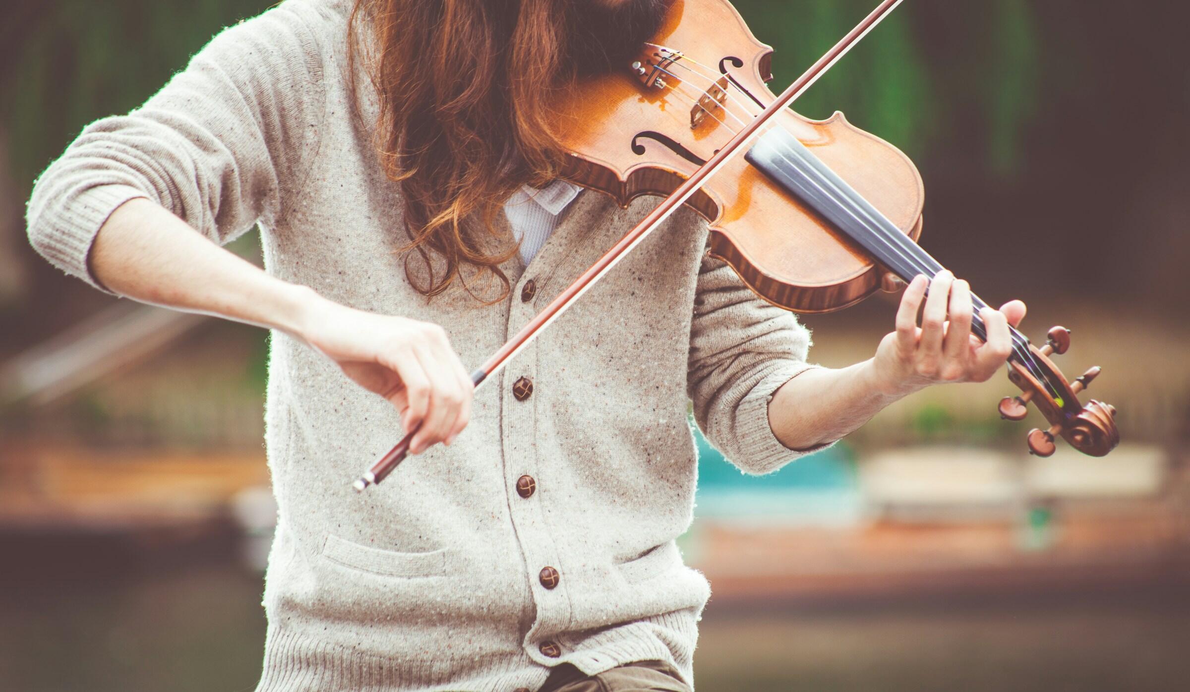Un músico con el pelo largo toca el violín al aire libre, vestido con un cómodo cárdigan beige, concentrado intensamente en su interpretación.