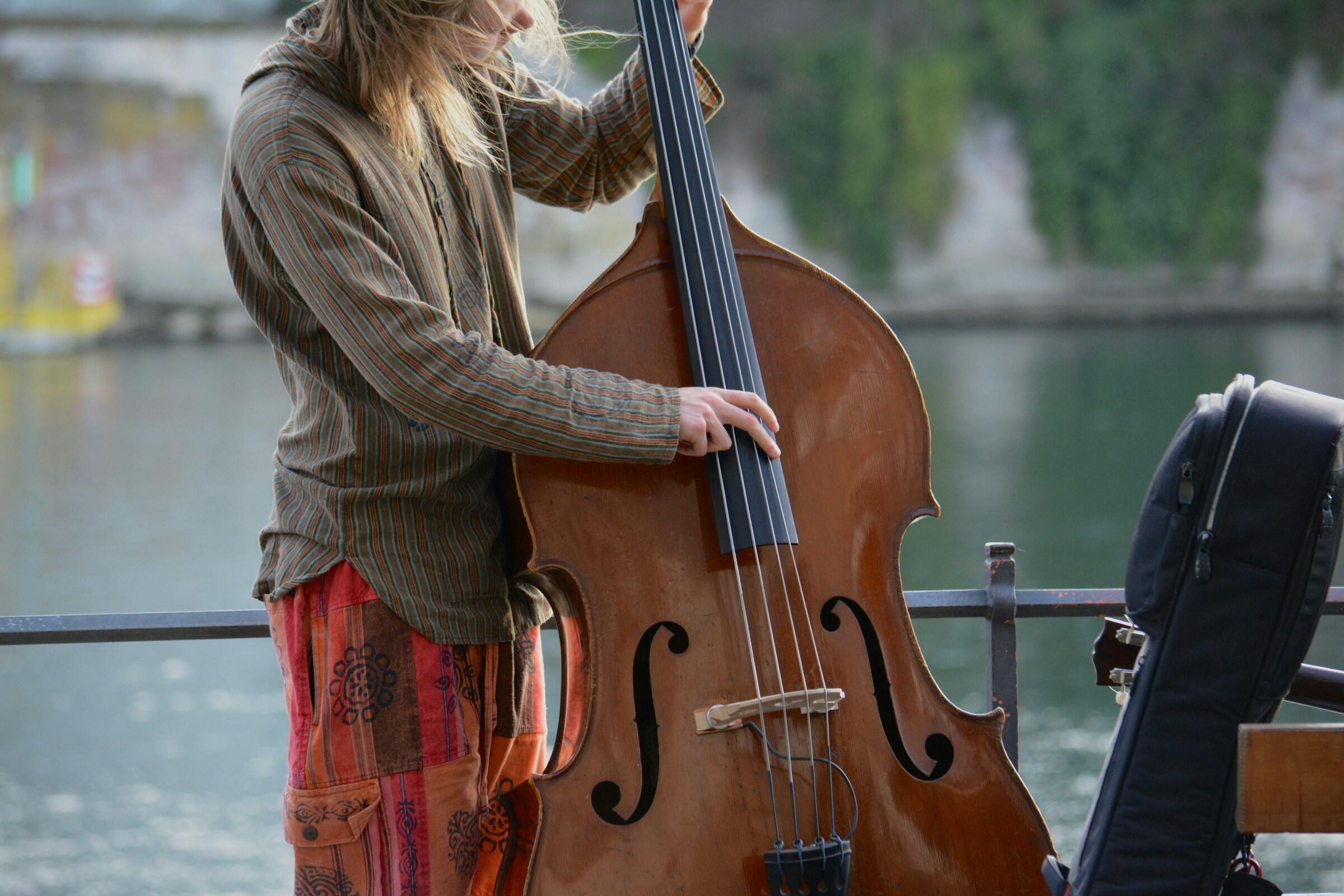 Un músico con una camisa a rayas toca el contrabajo junto al agua, con un estuche de guitarra cerca.