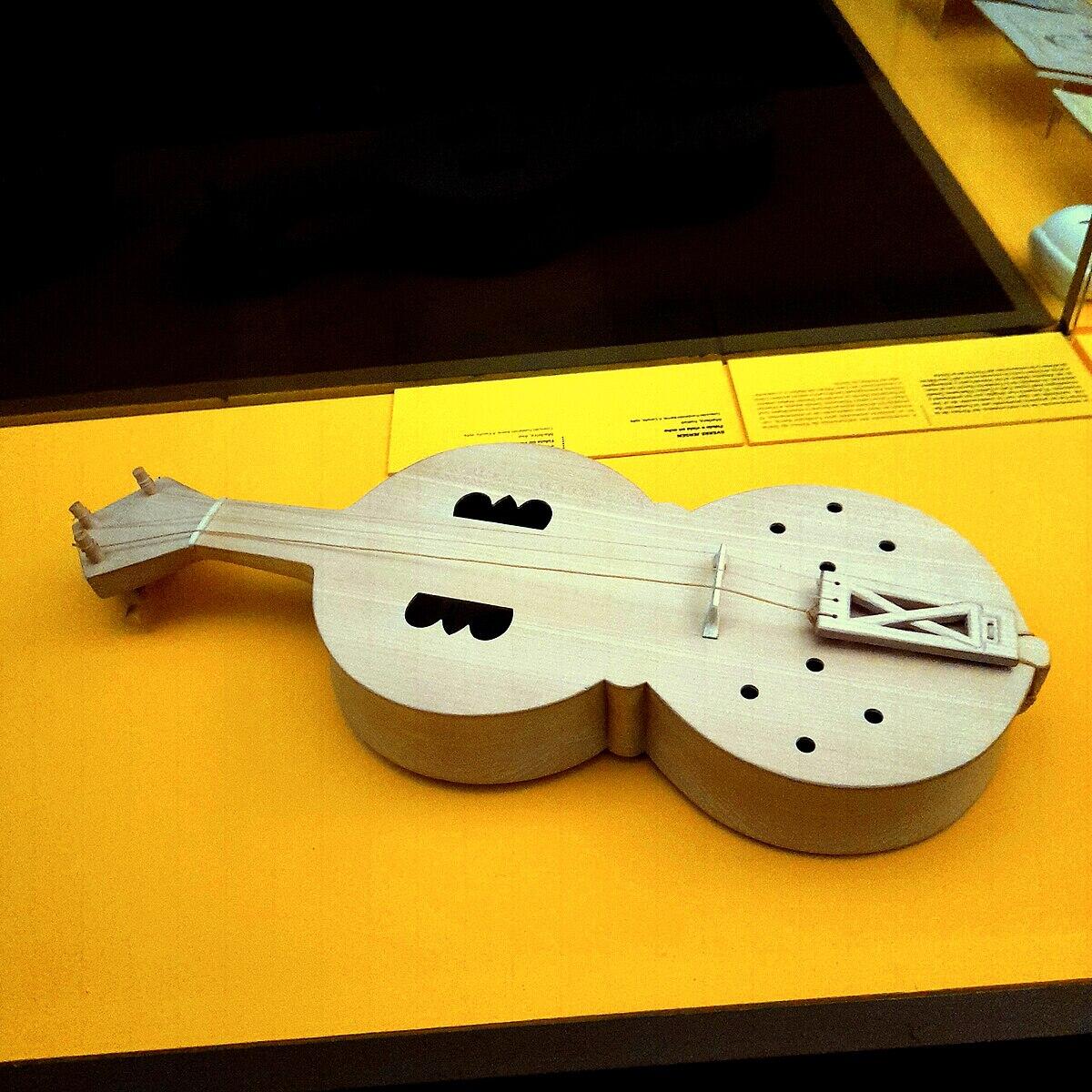 Instrumento de cuerda de madera con cuerpo redondeado y dos orificios acústicos, expuesto sobre una superficie amarilla brillante