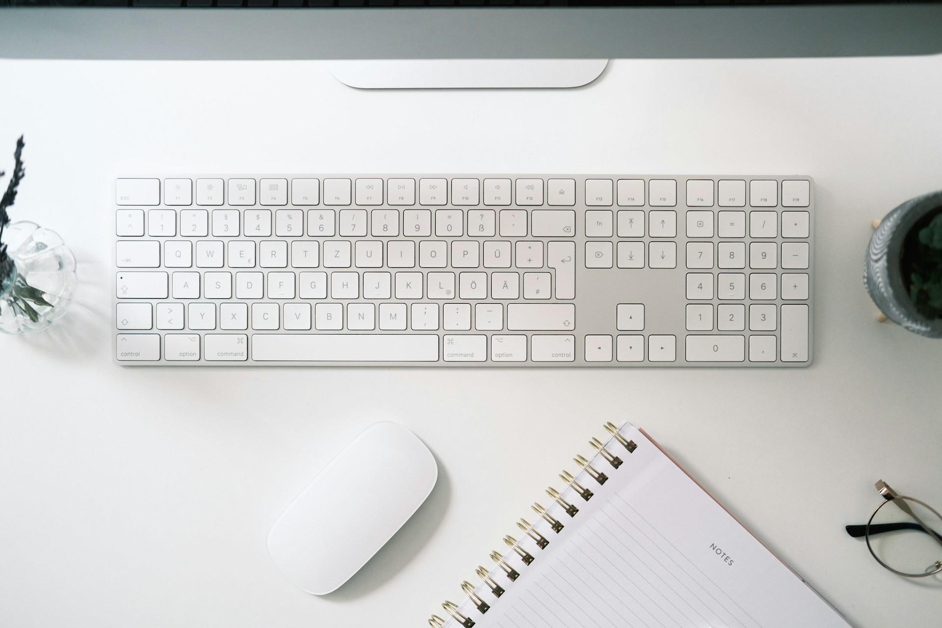 Un moderno teclado y ratón blancos sobre un escritorio con un cuaderno y una planta.