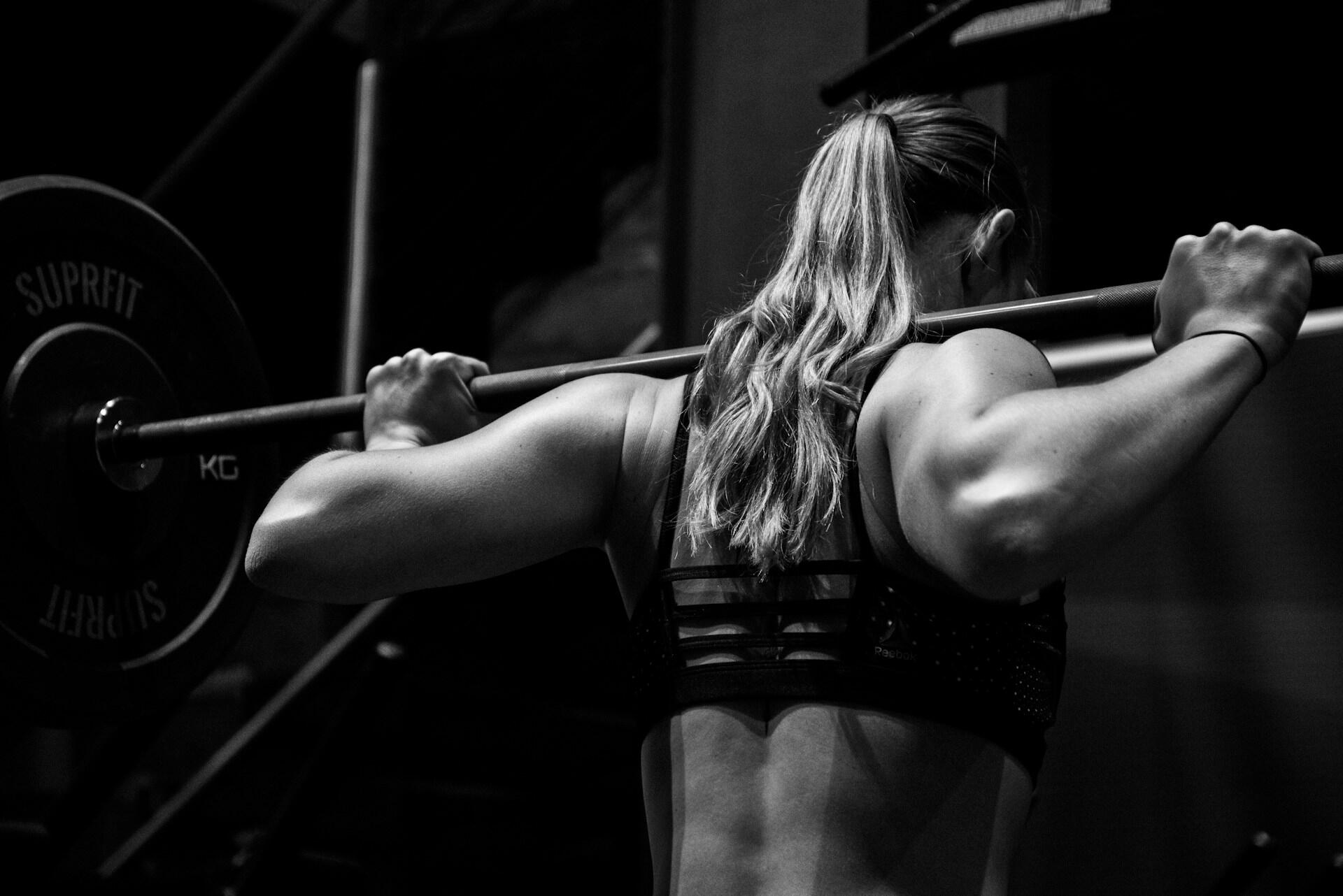 Una mujer de pelo largo, vista de espaldas, levanta una barra en un gimnasio. Lleva un sujetador deportivo y transmite fuerza y concentración en esta fotografía en blanco y negro.