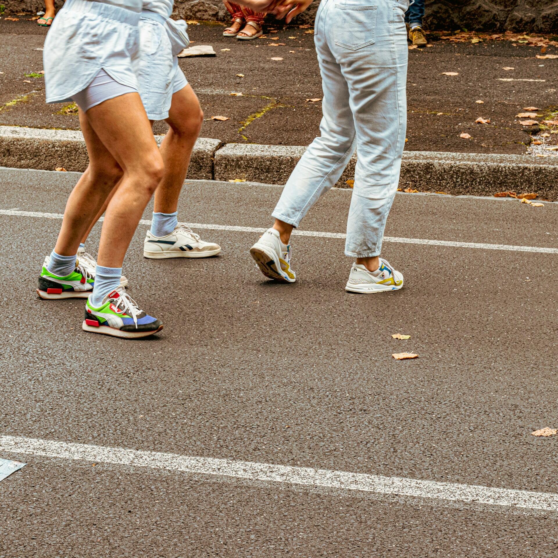 Gente con zapatillas de colores y ropa informal camina por una calle empedrada, con hojas esparcidas por el suelo. Se respira un ambiente otoñal y relajado.