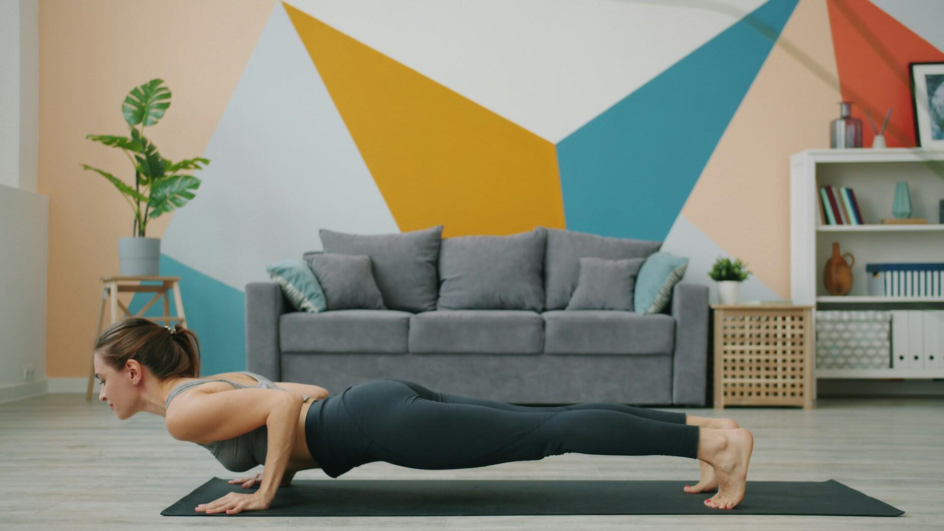 Una mujer practica yoga en el salón. Está haciendo la postura de la plancha sobre una esterilla negra, con una pared de motivos geométricos de colores, un sofá gris y plantas al fondo.