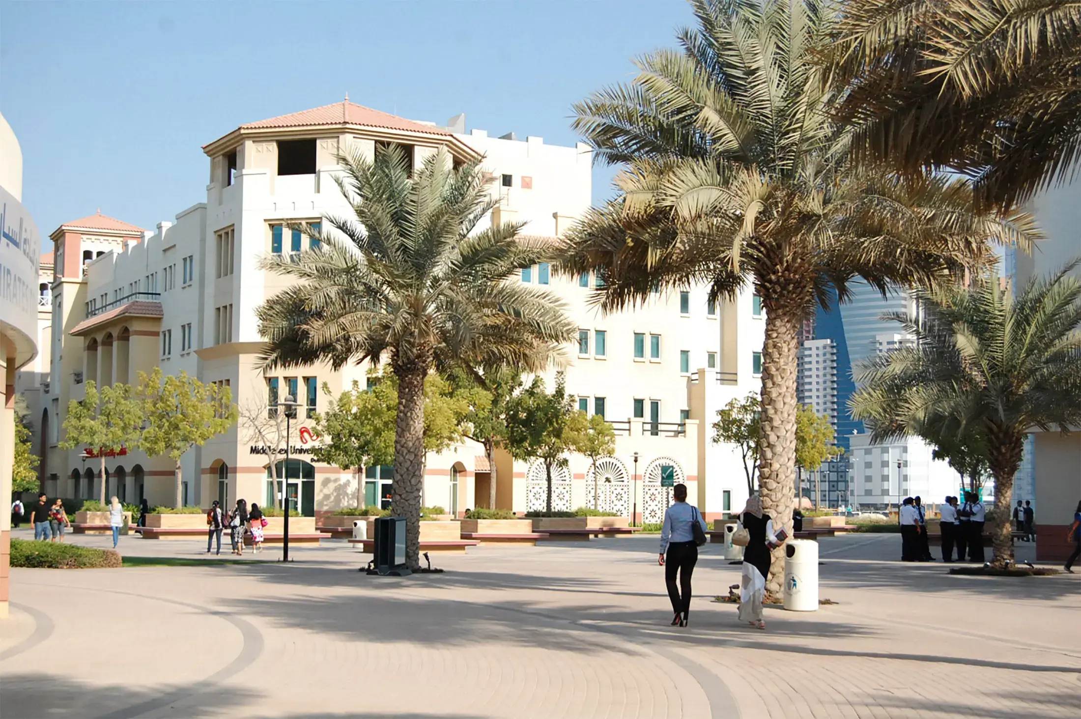 Una soleada escena en el campus con palmeras, edificios modernos y estudiantes caminando por un sendero pavimentado en una universidad.