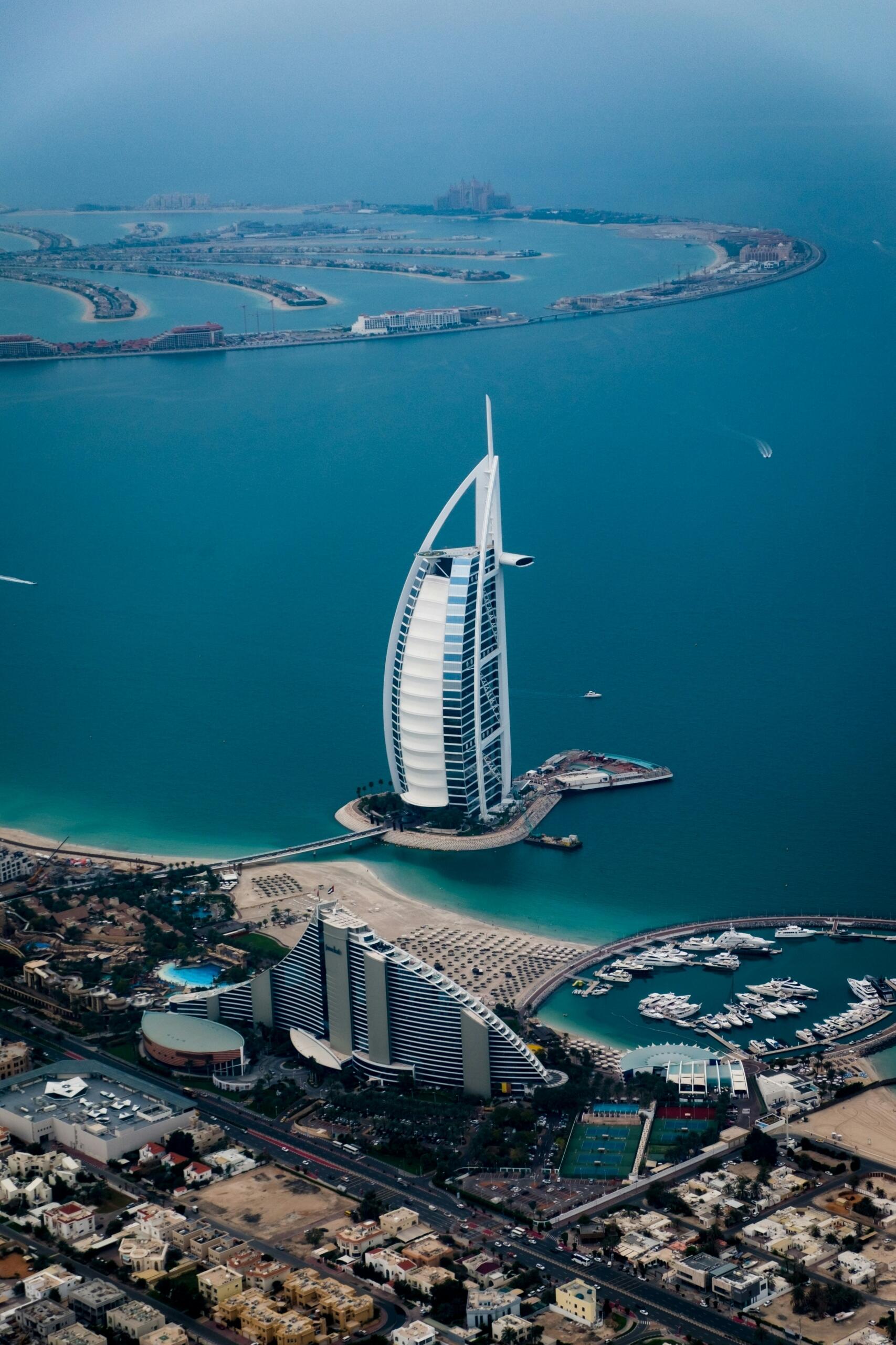 Vista aérea del emblemático hotel Burj Al Arab, rodeado de aguas turquesas y playas de arena, con lujosos barcos en el puerto deportivo.