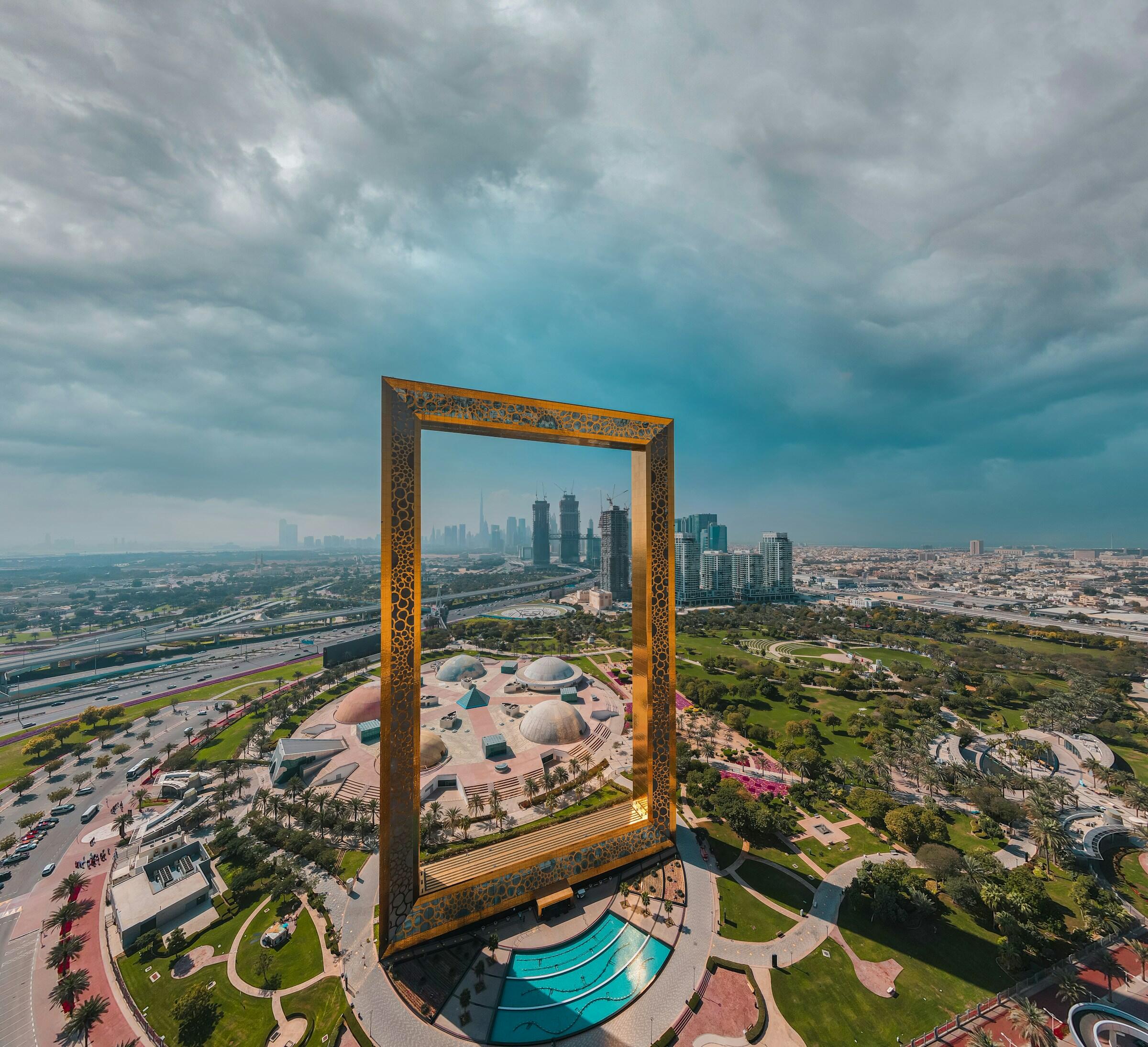 Vista aérea del Dubai Frame, una gran estructura dorada, con el paisaje urbano y los parques verdes visibles bajo un cielo nublado.