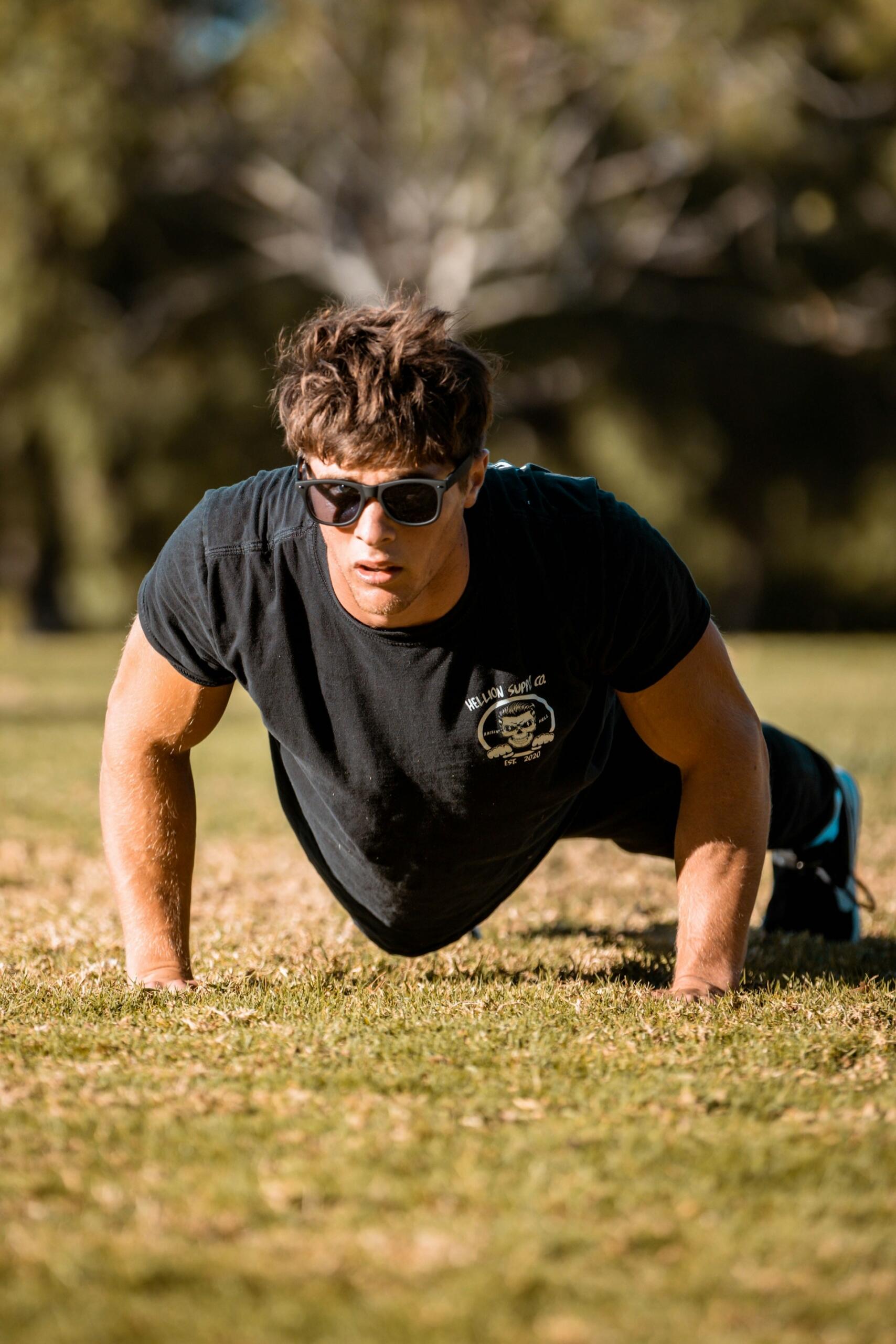 Un hombre con gafas de sol hace flexiones sobre el césped en un parque soleado. Viste una camiseta negra y parece concentrado y decidido, con árboles borrosos al fondo.