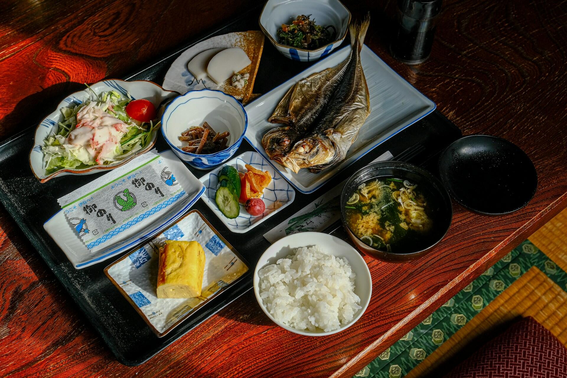 Una comida tradicional japonesa servida en una mesa de madera lacada: pescado a la parrilla, arroz, sopa de miso, ensalada, encurtidos y pequeños acompañamientos, todo ello con un aire acogedor y agradable.