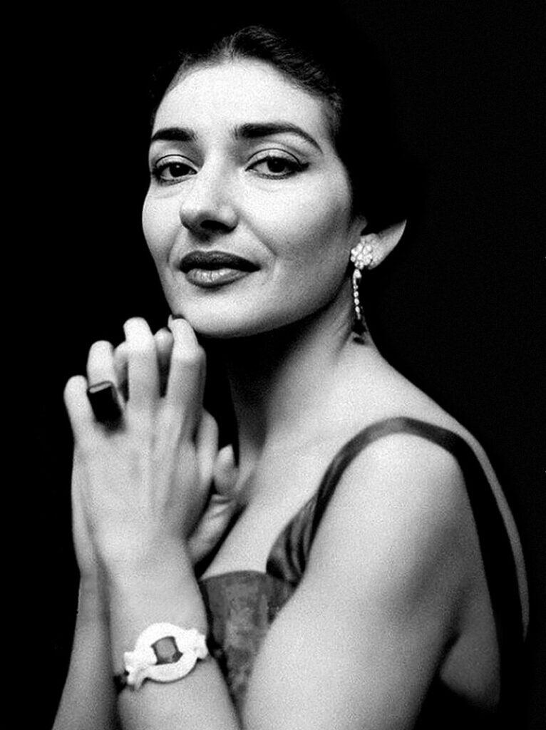 Retrato en blanco y negro de Maria Callas con una expresión elegante. Lleva pendientes, una pulsera y un anillo. Sus manos están entrelazadas con delicadeza, lo que transmite serenidad.