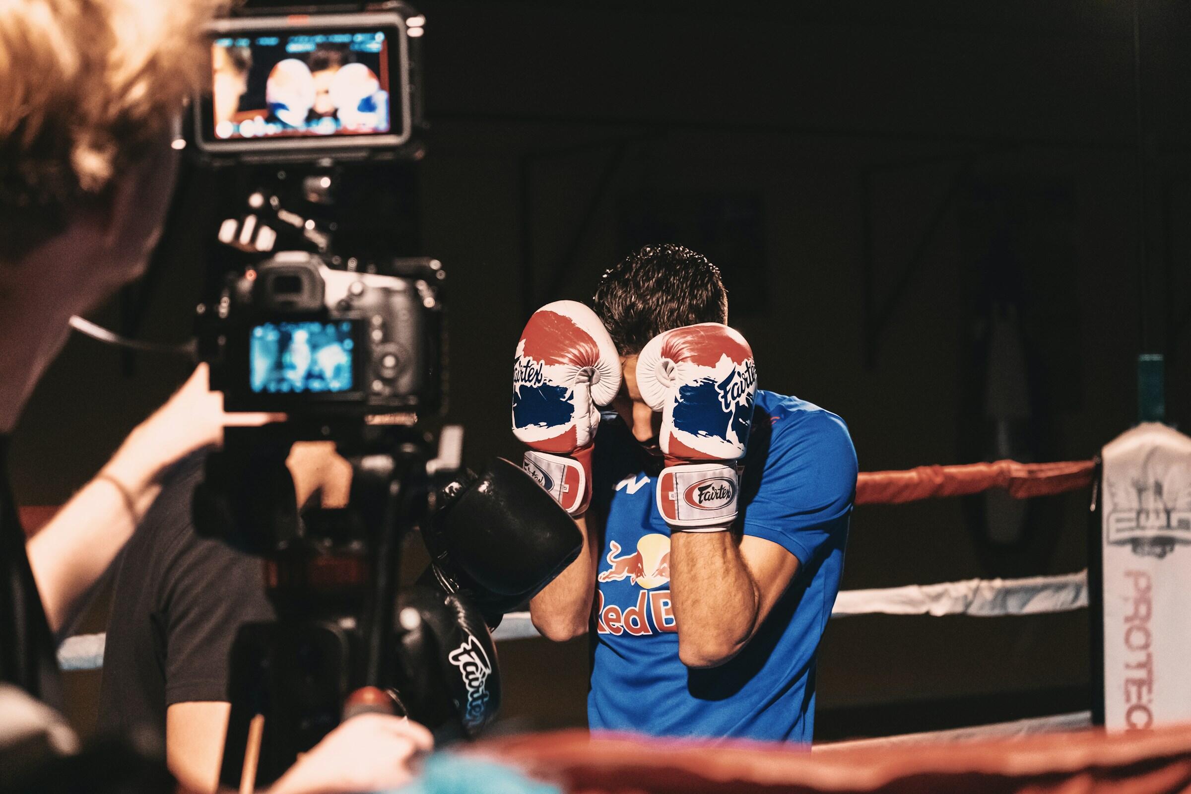 Un boxeador con camiseta azul y guantes rojos y blancos se protege instintivamente la cara, mientras una cámara captura el momento en un ring con poca luz.