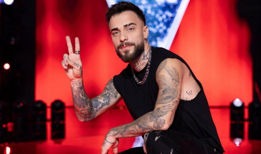 Diogo Piçarra tatuado se arrodilla en el escenario, sonriendo y haciendo el signo de la paz con los dedos. El fondo es rojo y vibrante, creando una atmósfera enérgica.