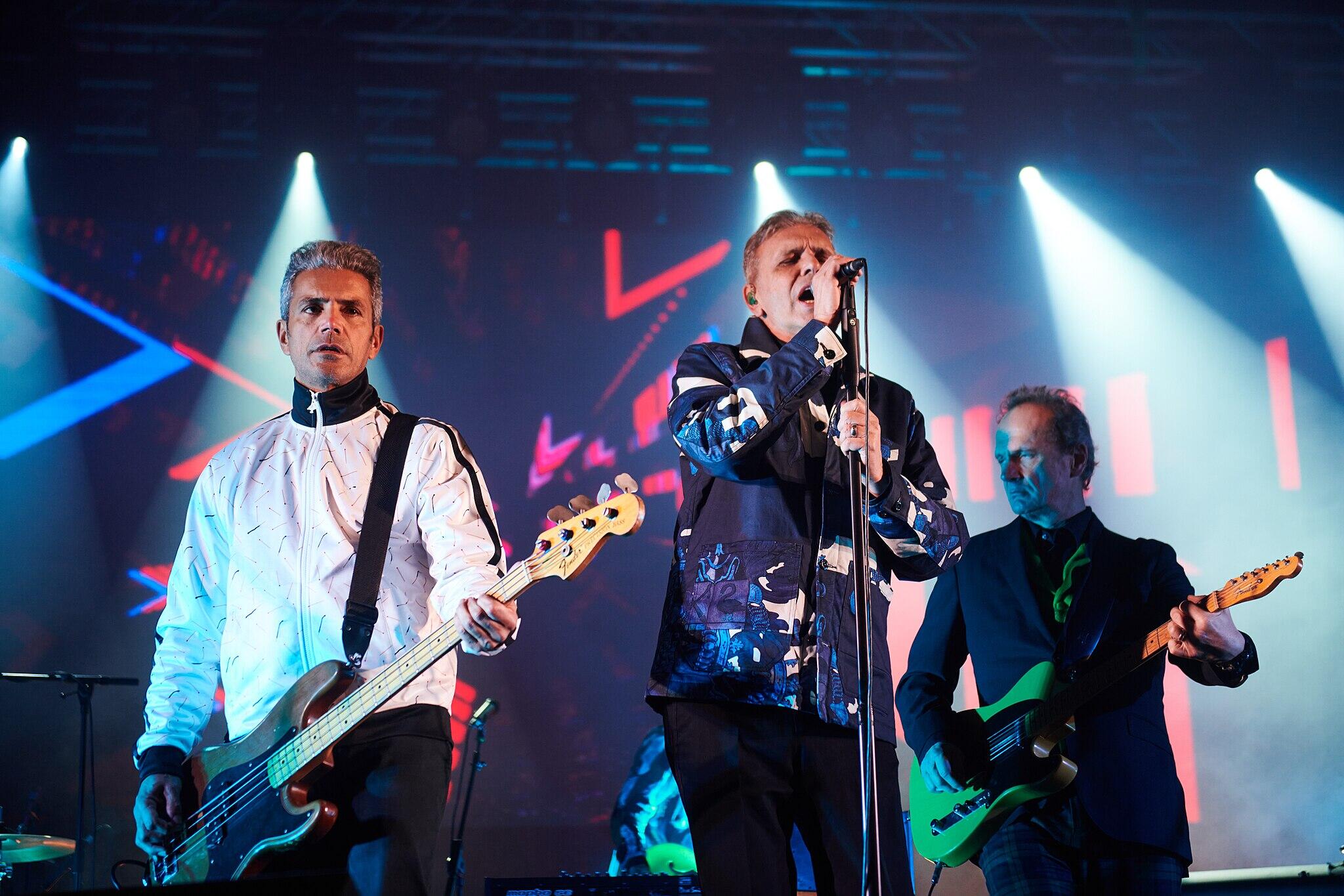 GNR en el escenario con luces vibrantes. El cantante principal, con una chaqueta estampada, canta al micrófono; dos guitarristas lo flanquean, creando una atmósfera enérgica.