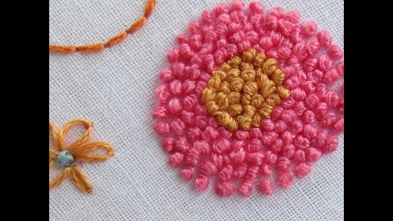 Primer plano de un bordado colorido que presenta una flor rosa con un centro dorado texturizado y una puntada decorativa naranja cerca.