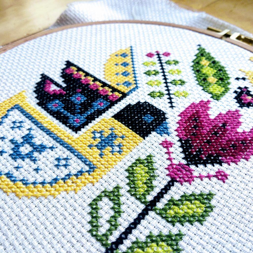 Bordado colorido con patrones florales y geométricos en negro, rosa, amarillo y verde cosidos sobre tela blanca.