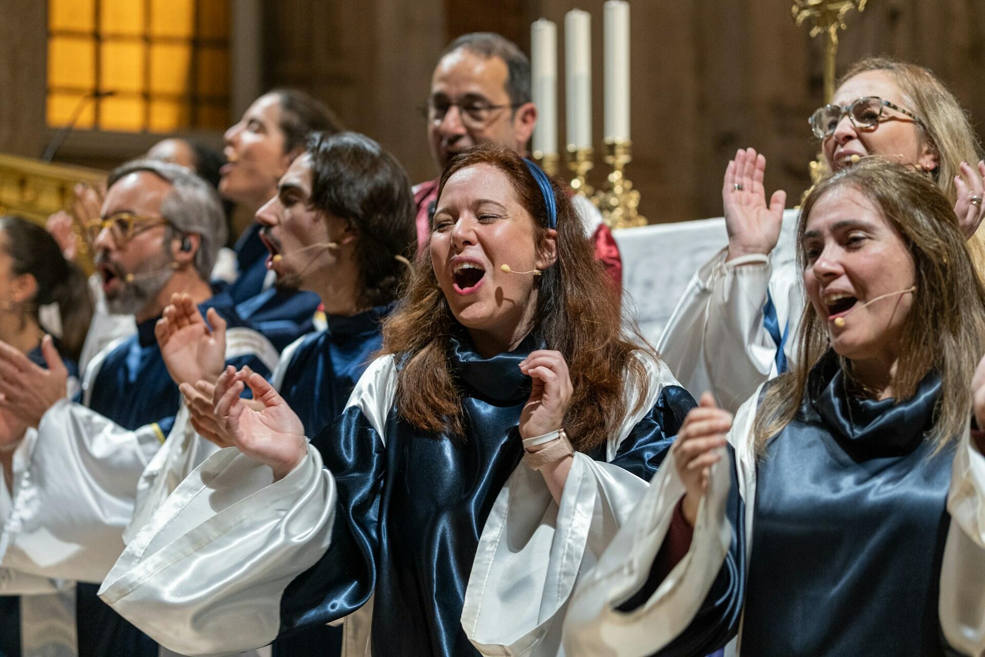 Un coro canta en una iglesia, con túnicas azules y blancas, y las manos alzadas expresando alegría y celebración. Se ven velas al fondo.
