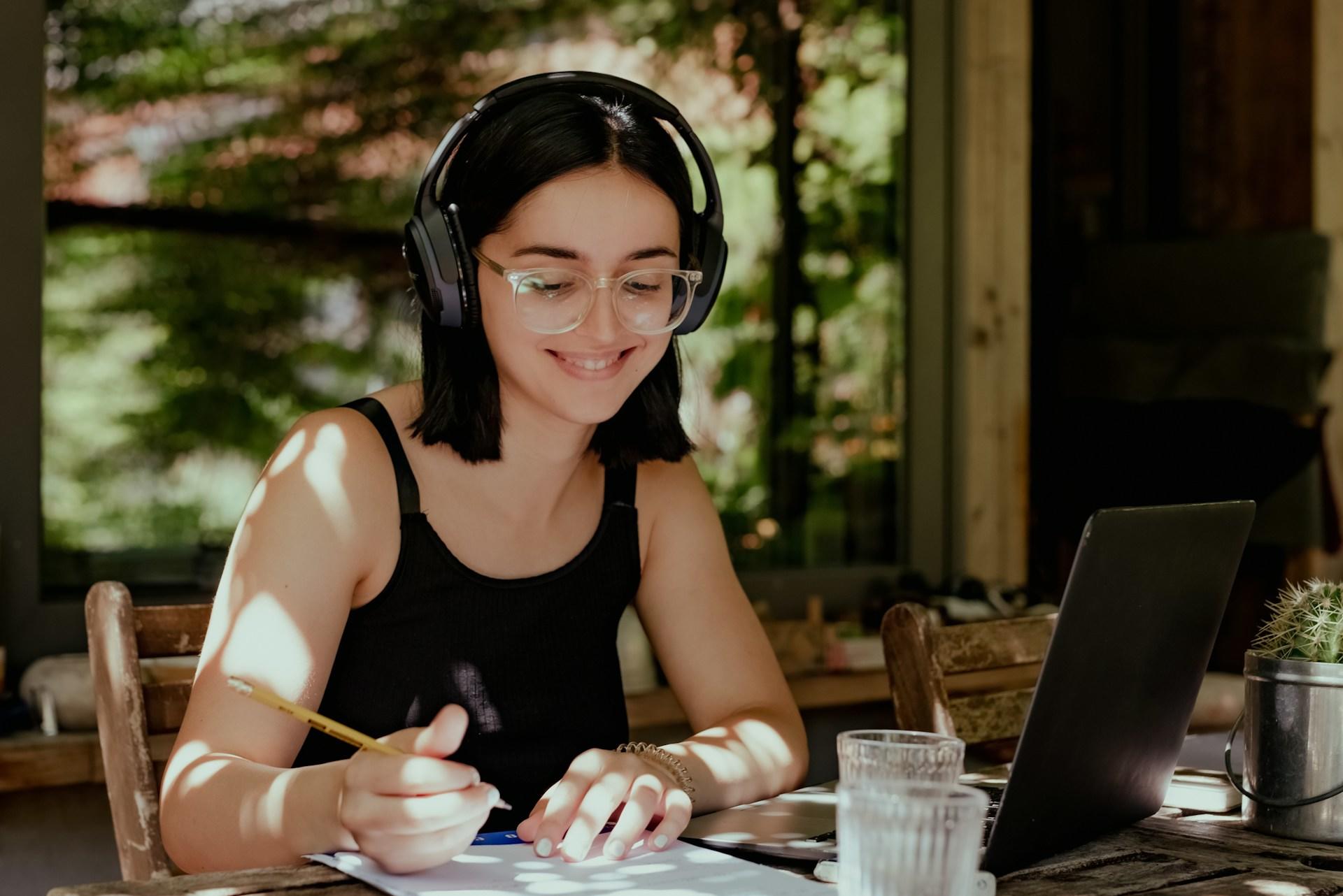 Una joven con auriculares sonríe mientras estudia en una mesa de madera. Escribe en un cuaderno junto a su ordenador portátil, rodeada de plantas y luz solar.