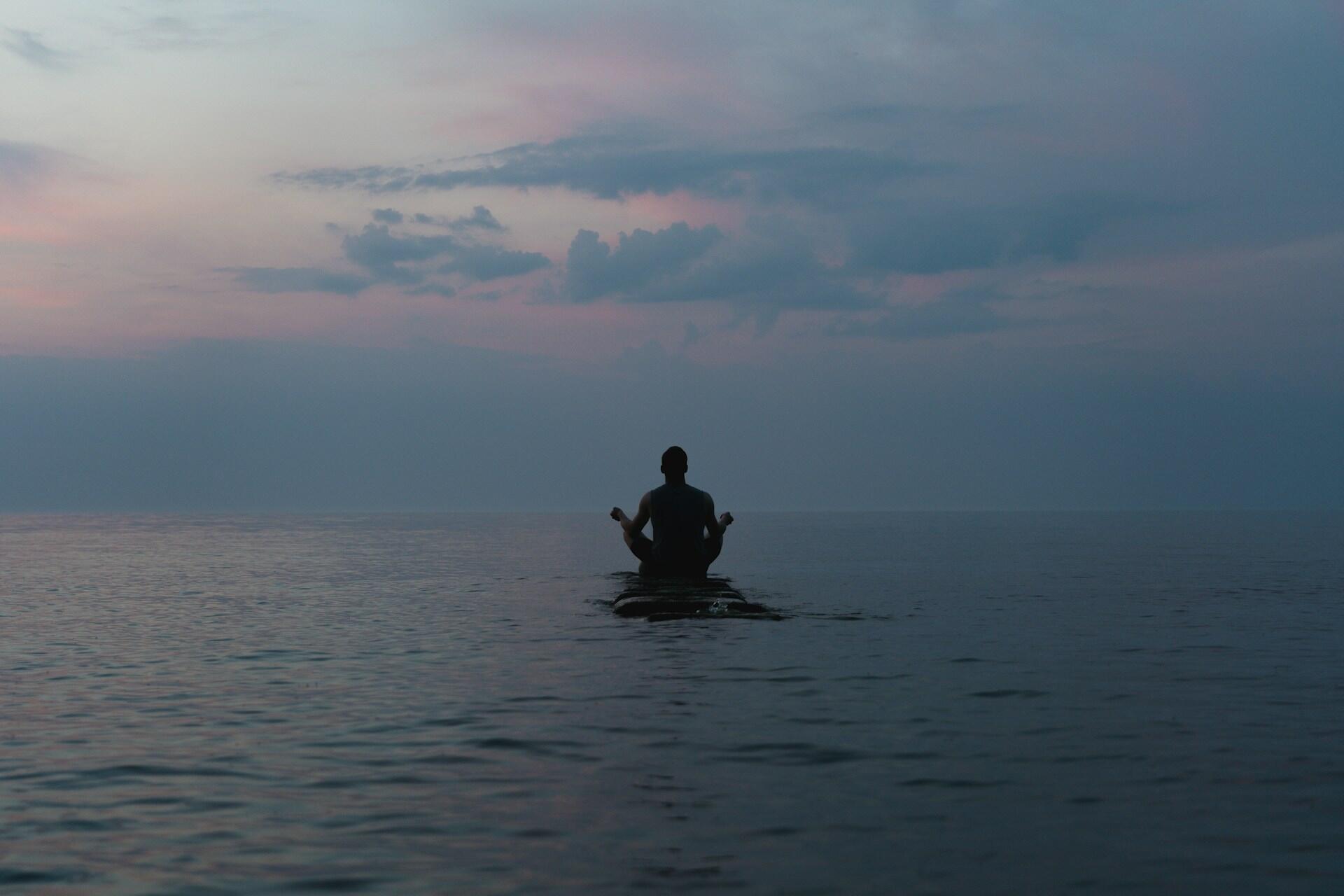 Una persona está sentada tranquilamente sobre una tabla de surf en el océano al atardecer, recortada contra un cielo sereno con suaves tonos rosados y azules, lo que transmite tranquilidad.