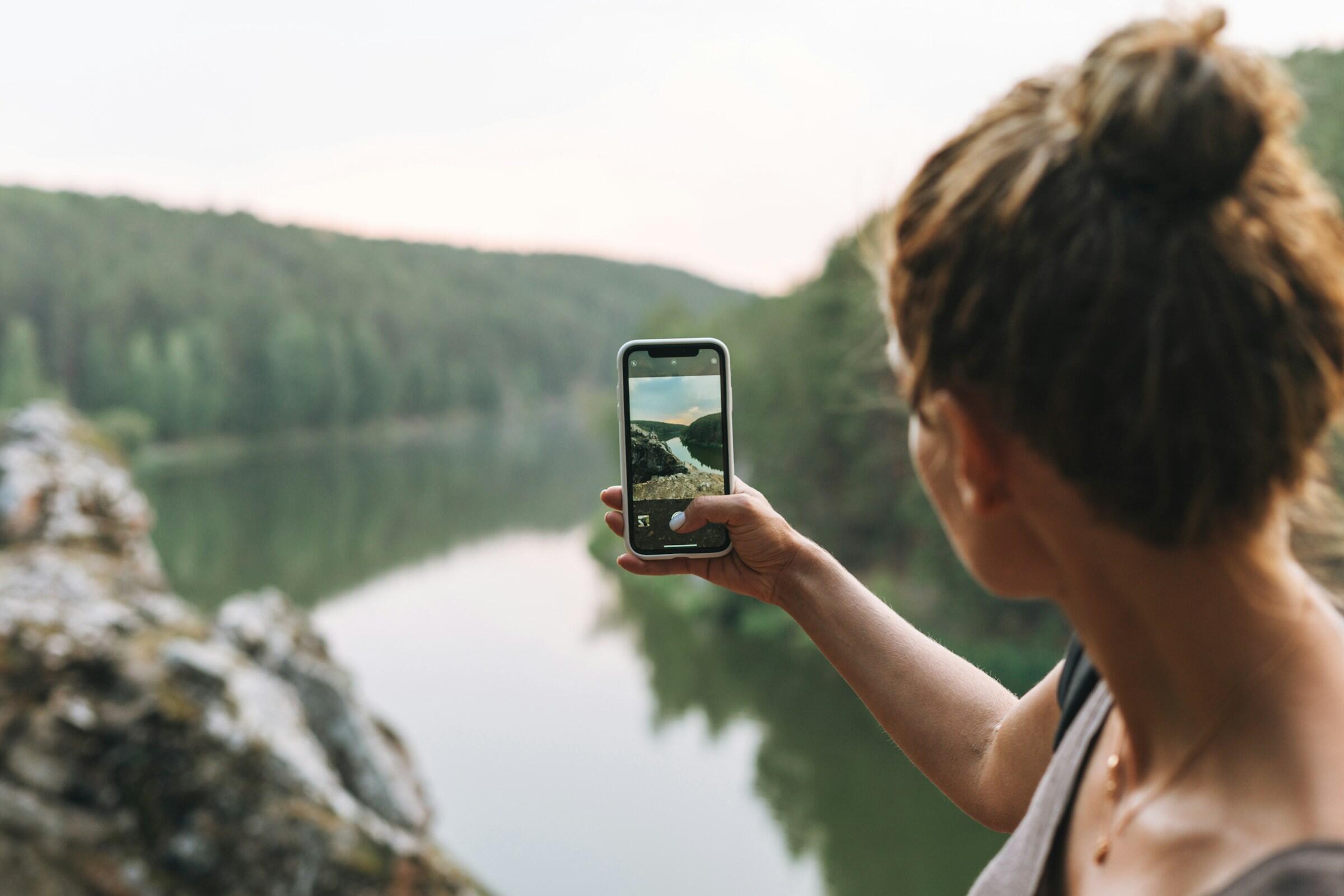 Una persona que fotografía un lago pintoresco y unas montañas boscosas con un smartphone.