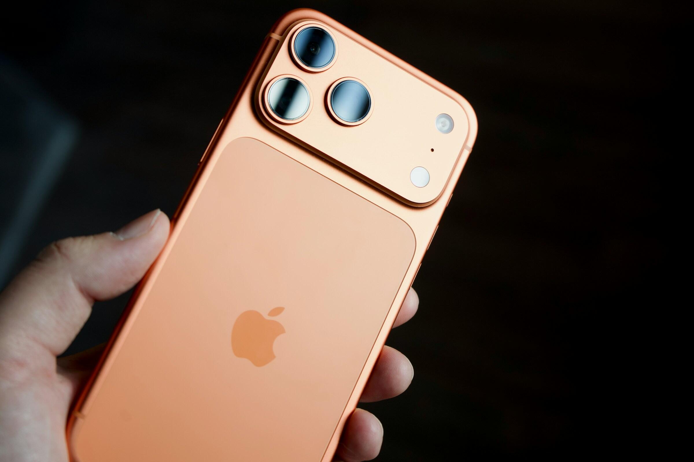Primer plano de un iPhone de color melocotón en la mano, en el que se aprecian el logotipo de Apple y el sistema de triple cámara trasera.