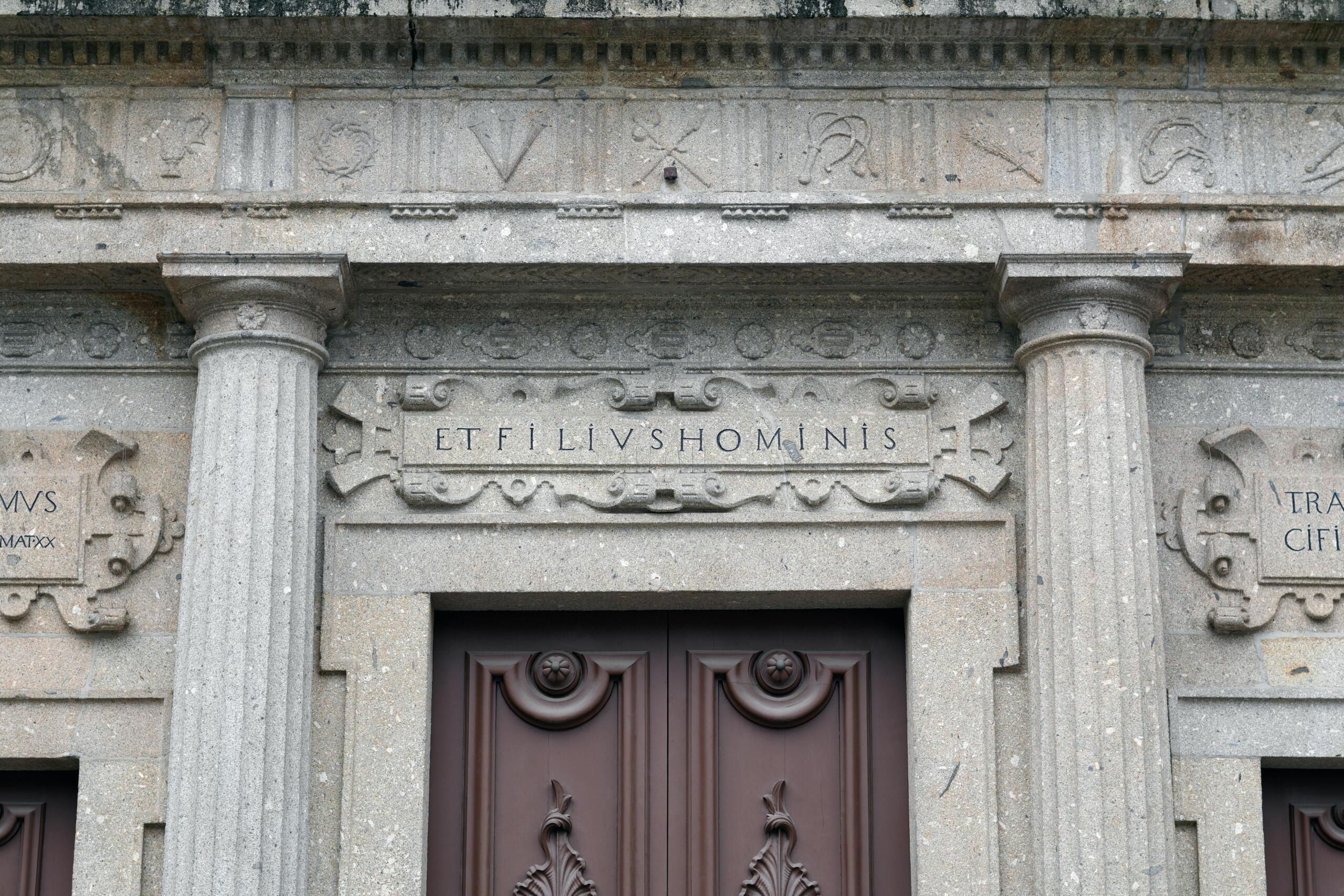 Detalle arquitectónico de una fachada de piedra clásica con columnas estriadas, una puerta de madera oscura y una inscripción central tallada en latín que reza 'ET FILIVS HOMINIS', bajo un friso decorativo con símbolos tallados.