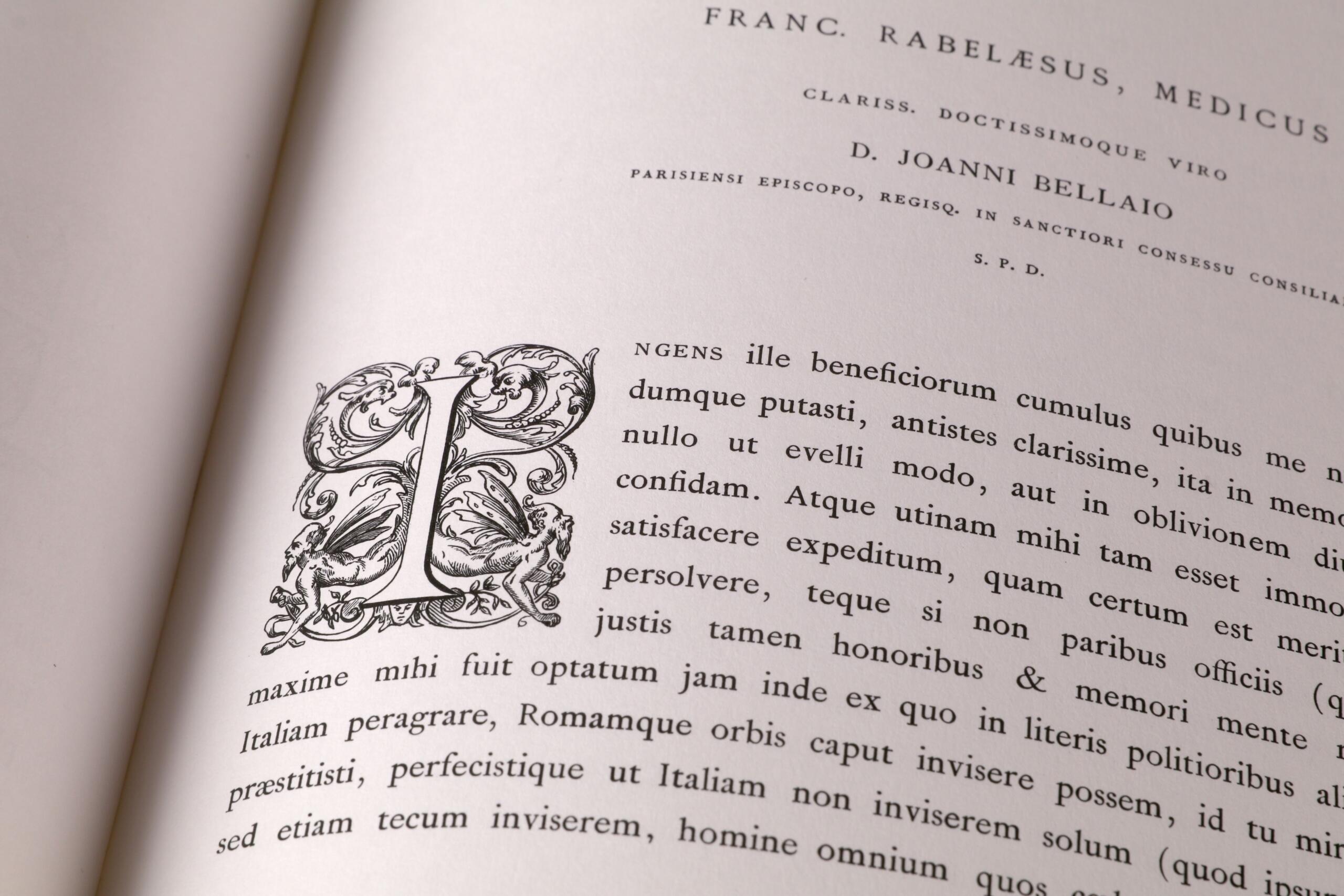 Primer plano de una página de un libro en la que se ve una «I» mayúscula ornamentada con un texto en latín a su lado, lo que pone de relieve la tipografía clásica.