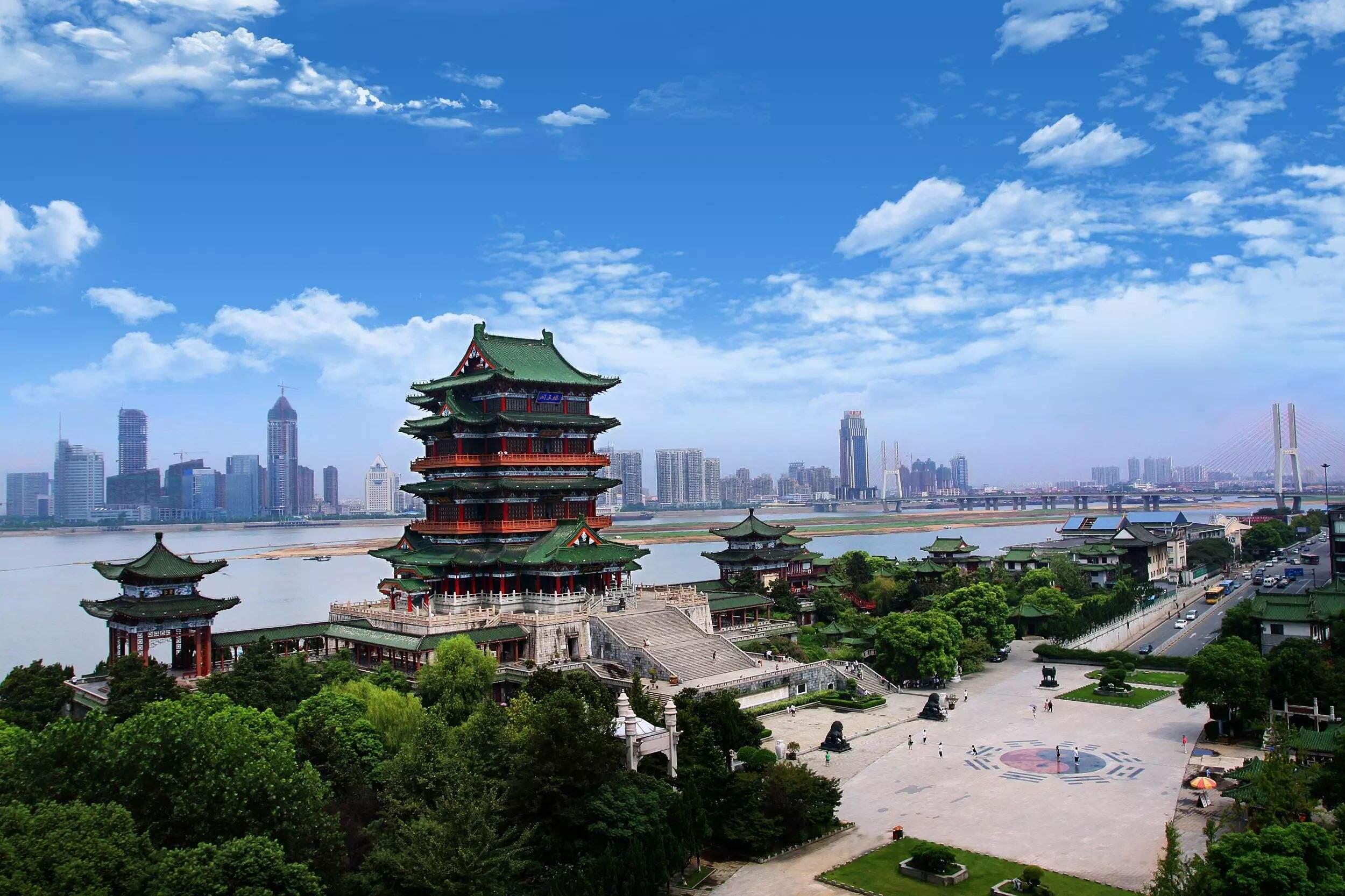 Una pagoda de arquitectura tradicional china, en tonos verdes y rojos, se eleva sobre unos exuberantes jardines. Al otro lado del río, bajo un cielo azul brillante, se divisa el perfil urbano de una ciudad moderna.