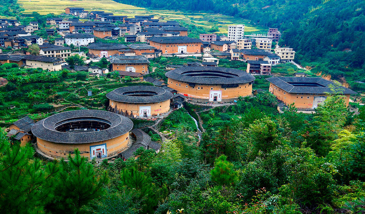 Vista aérea de los Fujian Tulou, construcciones tradicionales chinas circulares de adobe, rodeadas de una exuberante vegetación y pequeñas casas de pueblo en un tranquilo entorno rural.