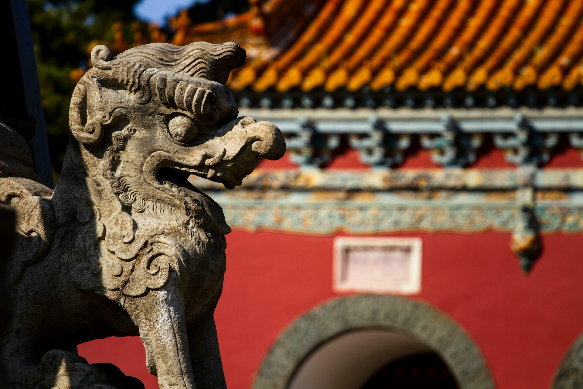 Una estatua de león de piedra con detalles intrincados en primer plano, con el telón de fondo de la pared de un templo de un rojo intenso y un tejado naranja ornamentado, que transmite una sensación de cultura ancestral.
