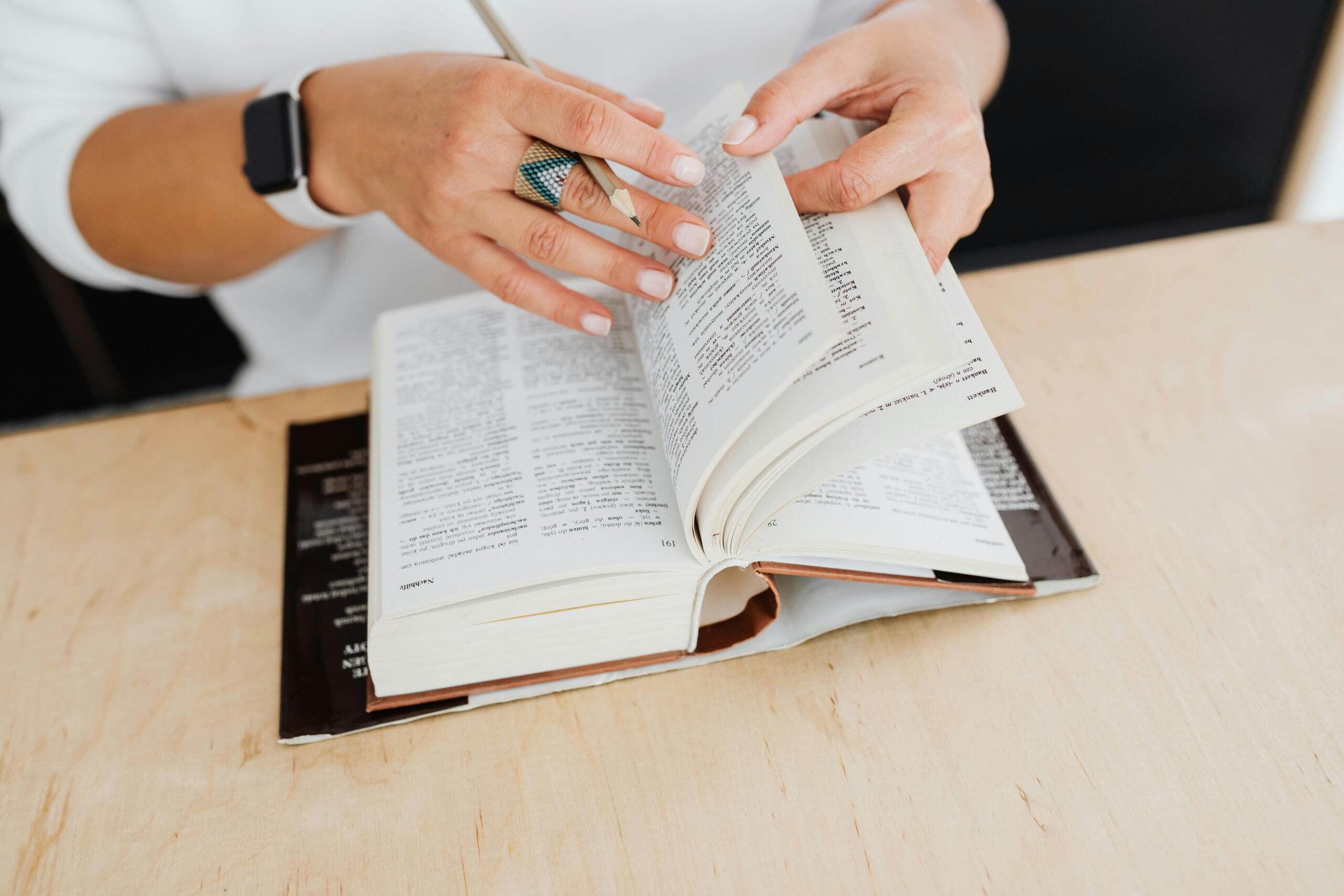 Una persona que hojea un libro grueso con una mano y sostiene un lápiz con la otra, sentada ante una mesa de madera.