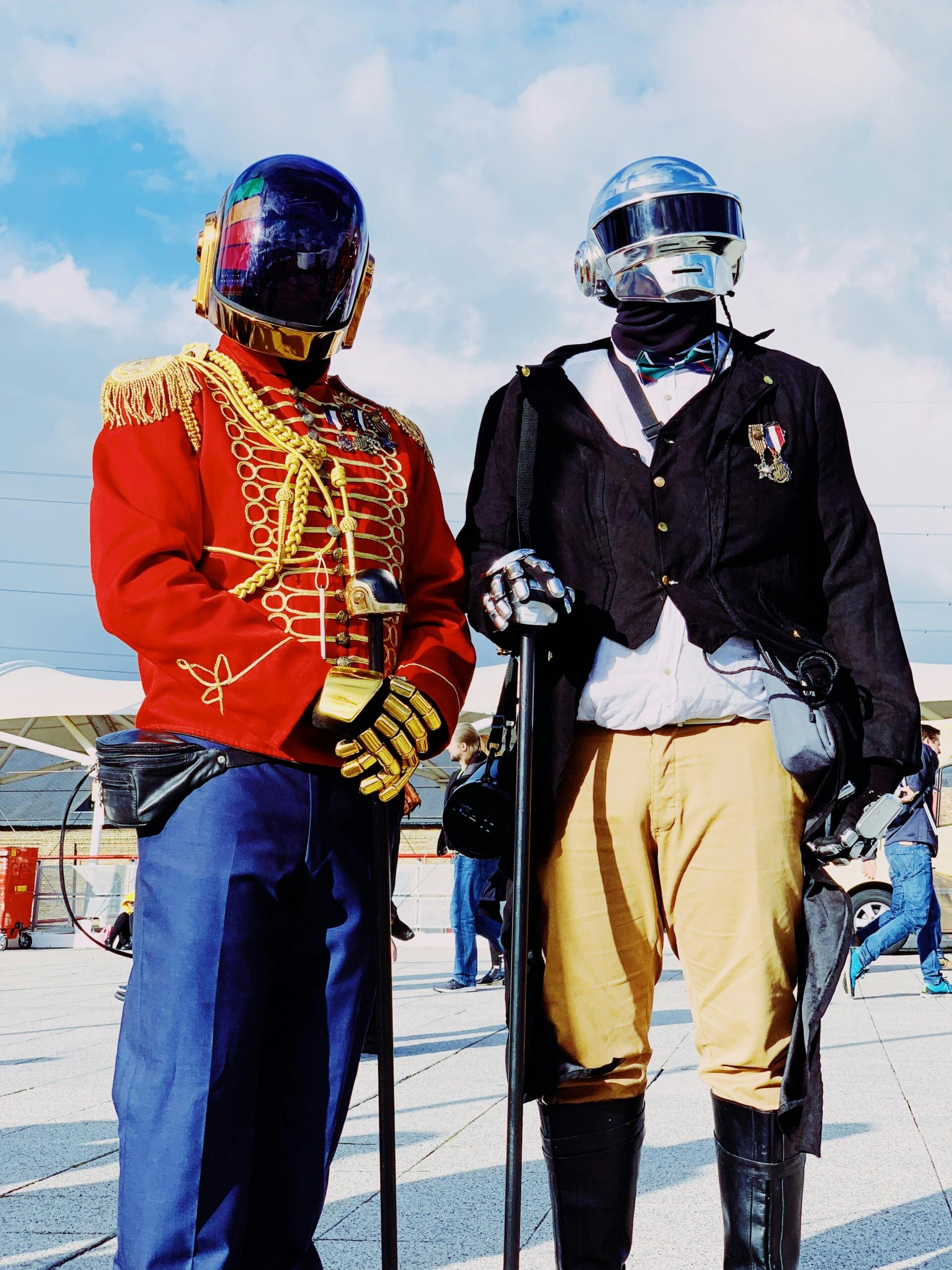 Dos personas con elaborados trajes robóticos parecidos a los de Daft Punk están juntas, una con una chaqueta militar roja y la otra con un esmoquin.