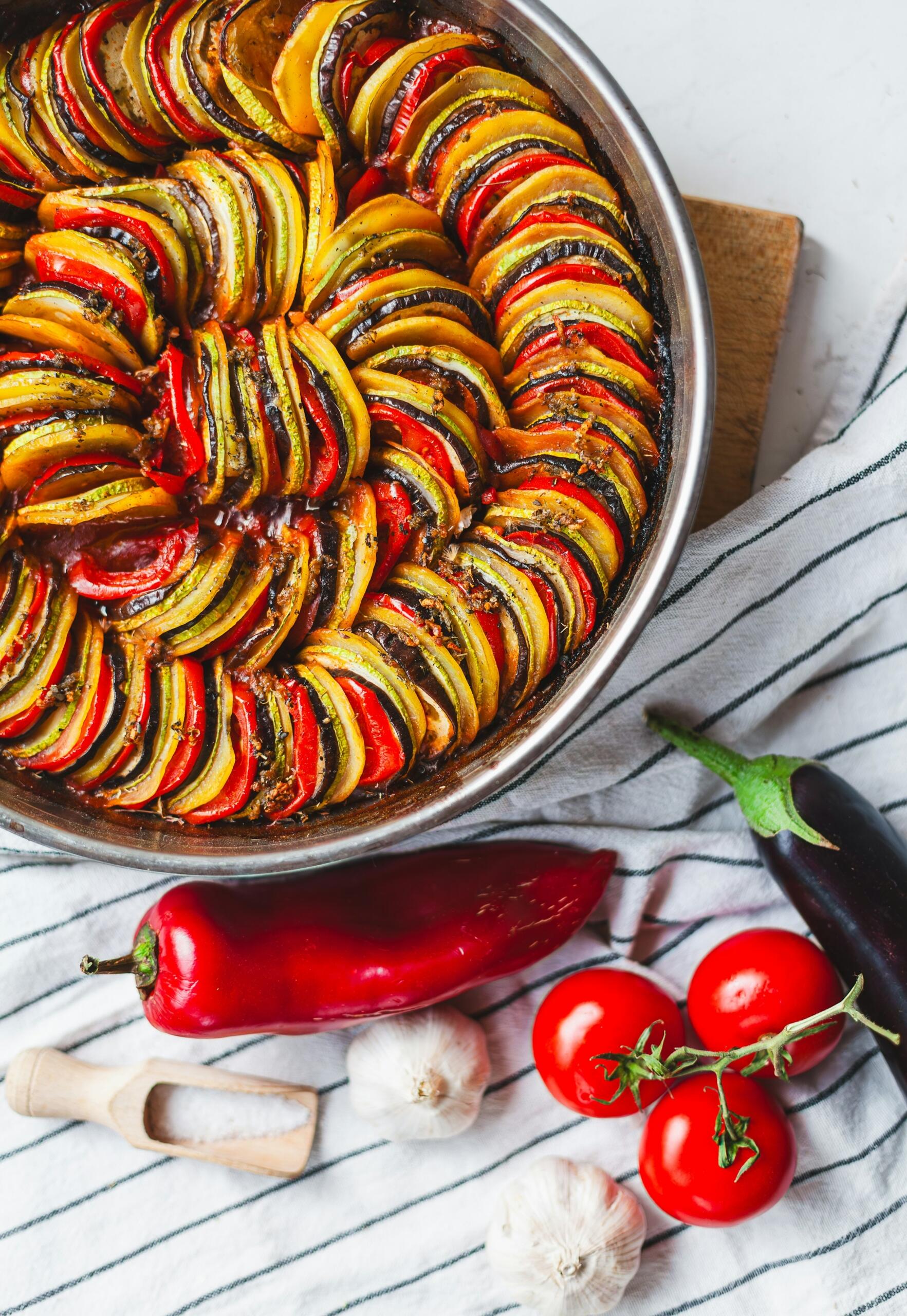 Un colorido plato de pisto en una sartén plateada, rodeado de verduras frescas, ajo y una cuchara de madera para la sal sobre un mantel a rayas.