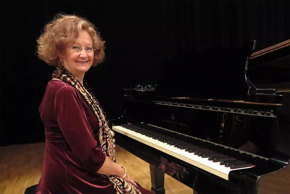 la pianista española Ángeles López Artiga con un vestido burdeos y un pañuelo estampado está sentada junto a un piano de cola en una habitación con poca luz.