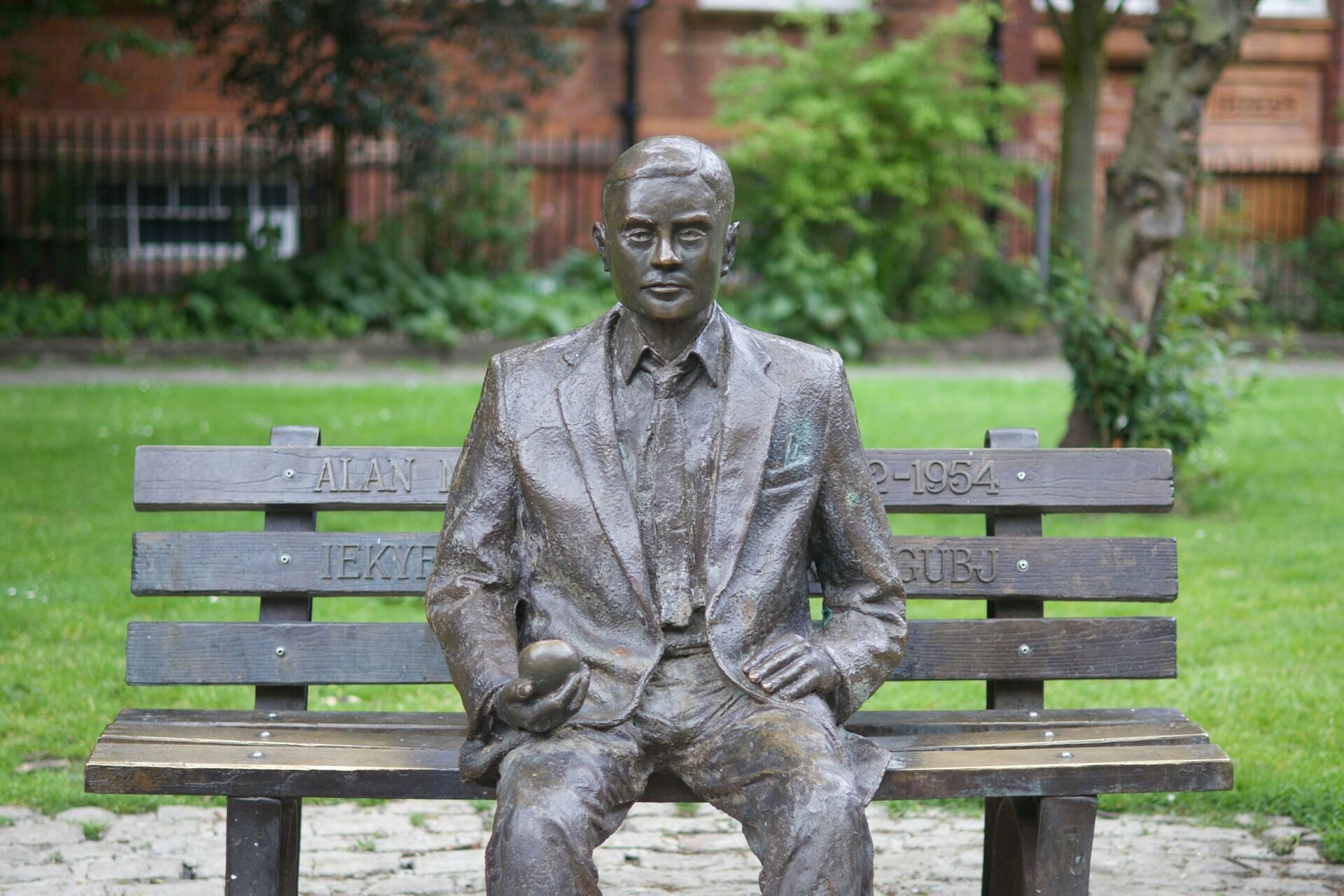 Estatua de bronce de Alan Turing sentado en un banco de madera en un parque, sosteniendo una manzana. La exuberante vegetación y un edificio de ladrillo en el fondo borroso transmiten una atmósfera serena.