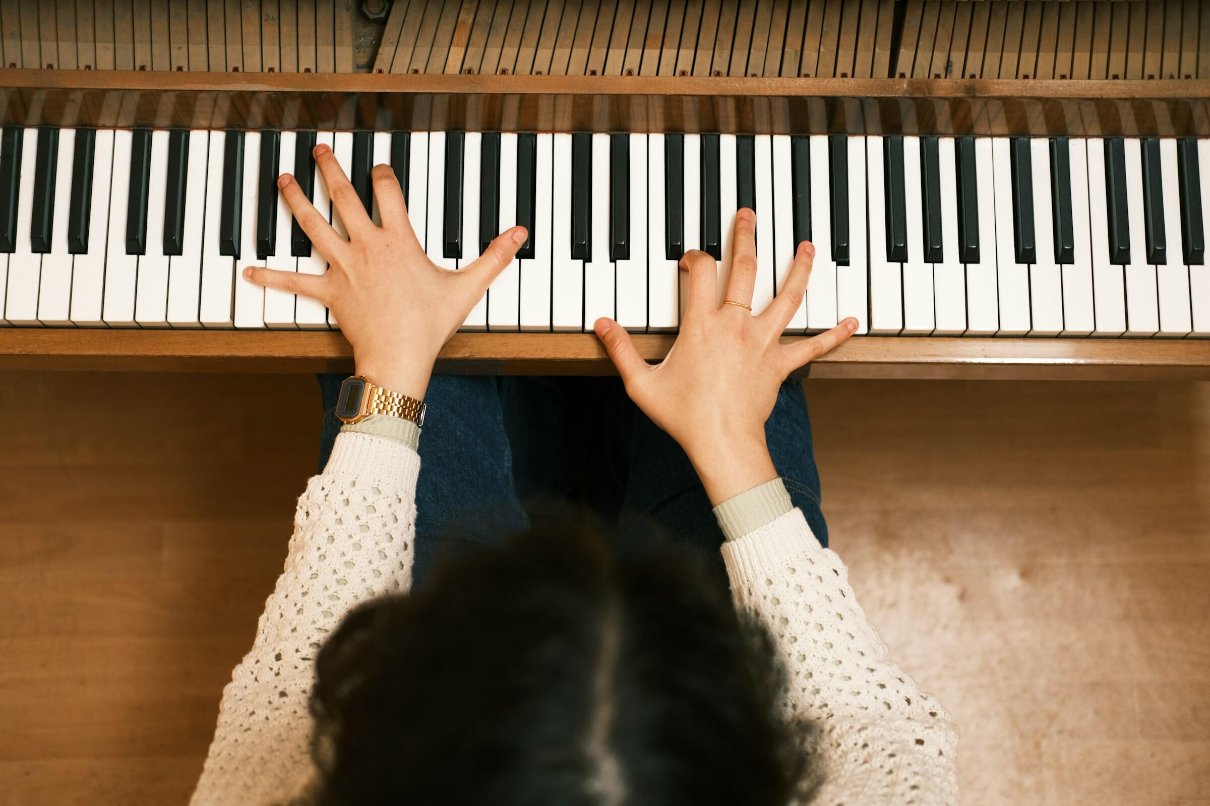Vista aérea de unas manos tocando el piano, una mano sobre las teclas negras y la otra sobre las teclas blancas, con un reloj y un anillo.