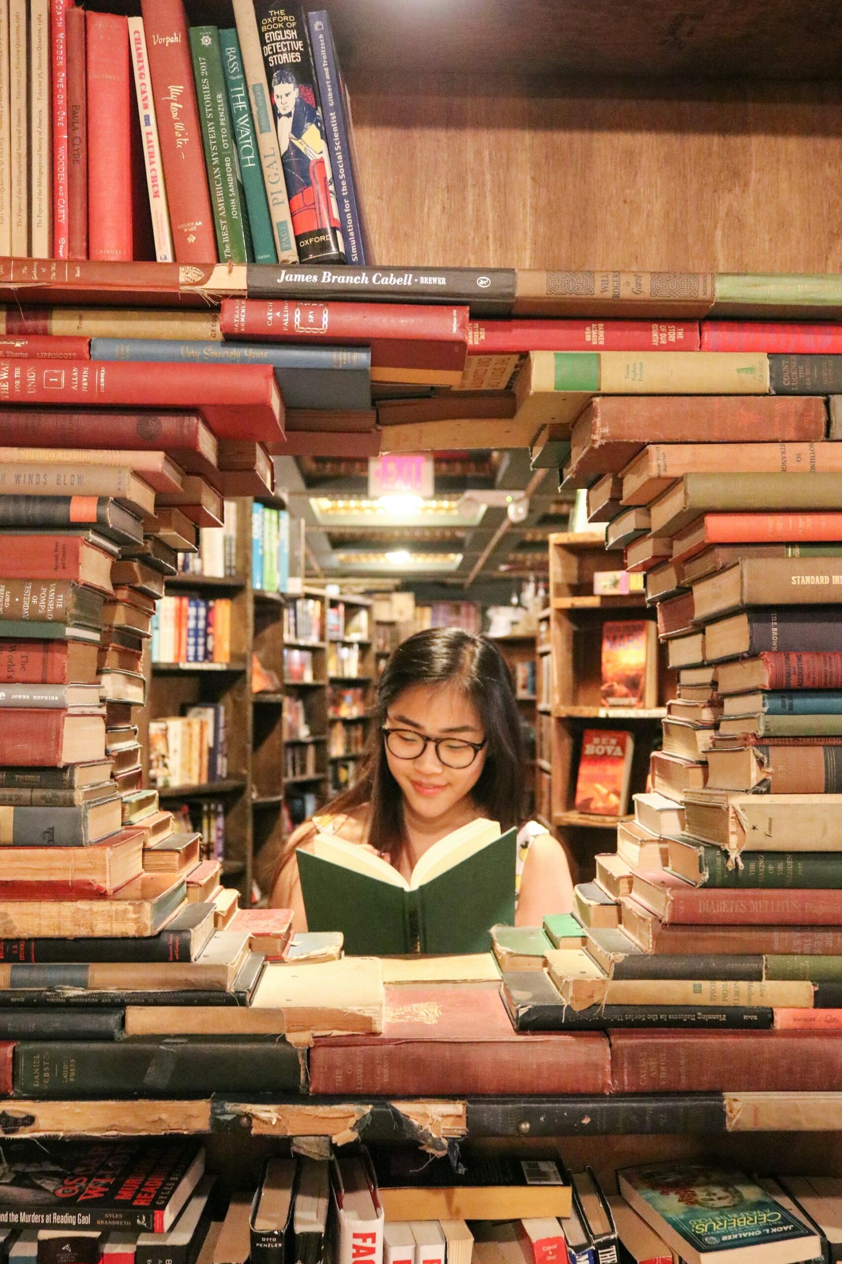 Una mujer está sentada en una biblioteca, absorta en la lectura de un libro, rodeada de estanterías llenas de libros.