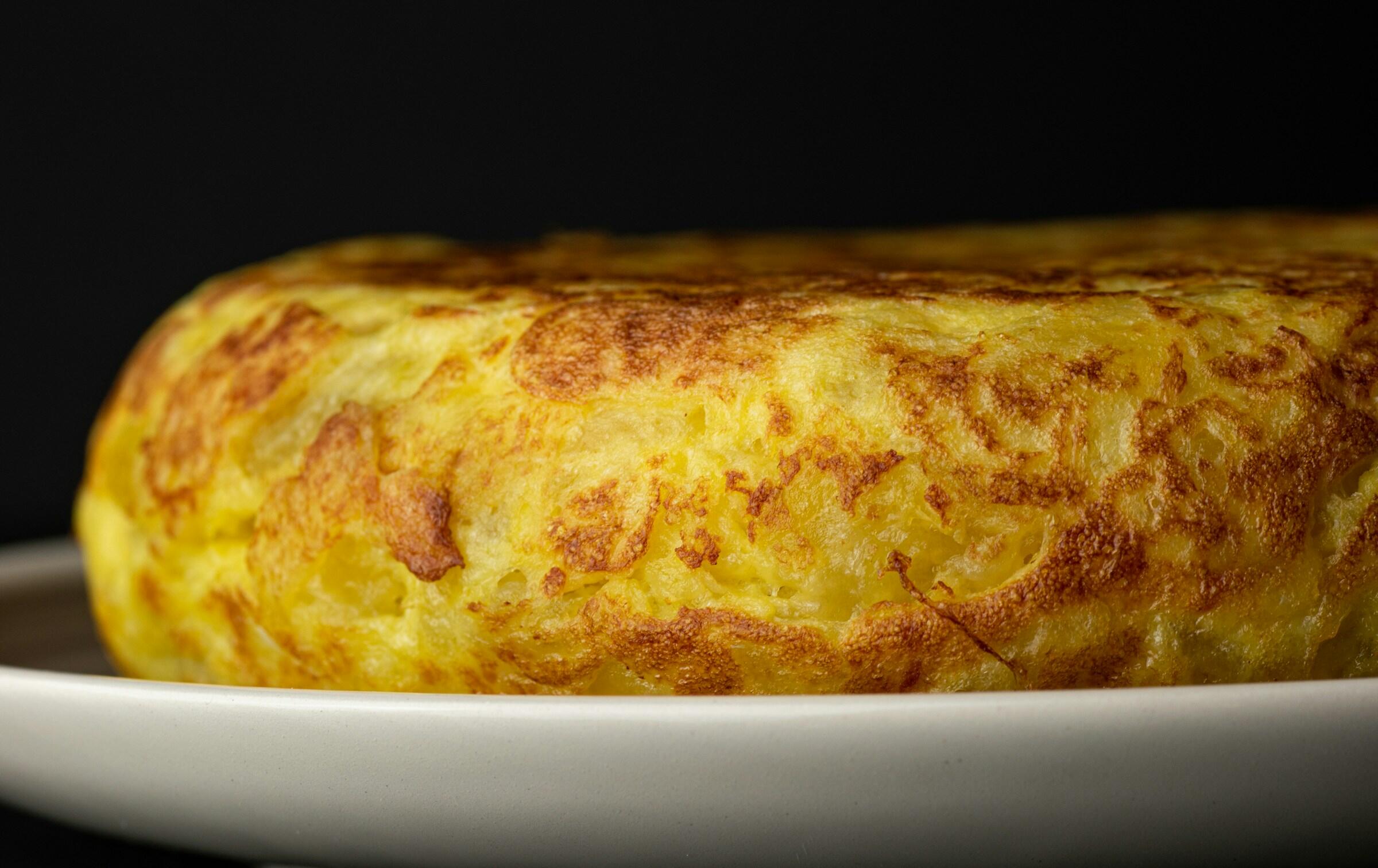 Primer plano de una tortilla española dorada sobre un plato blanco, en el que se aprecia su textura esponjosa y su exterior crujiente sobre un fondo oscuro.