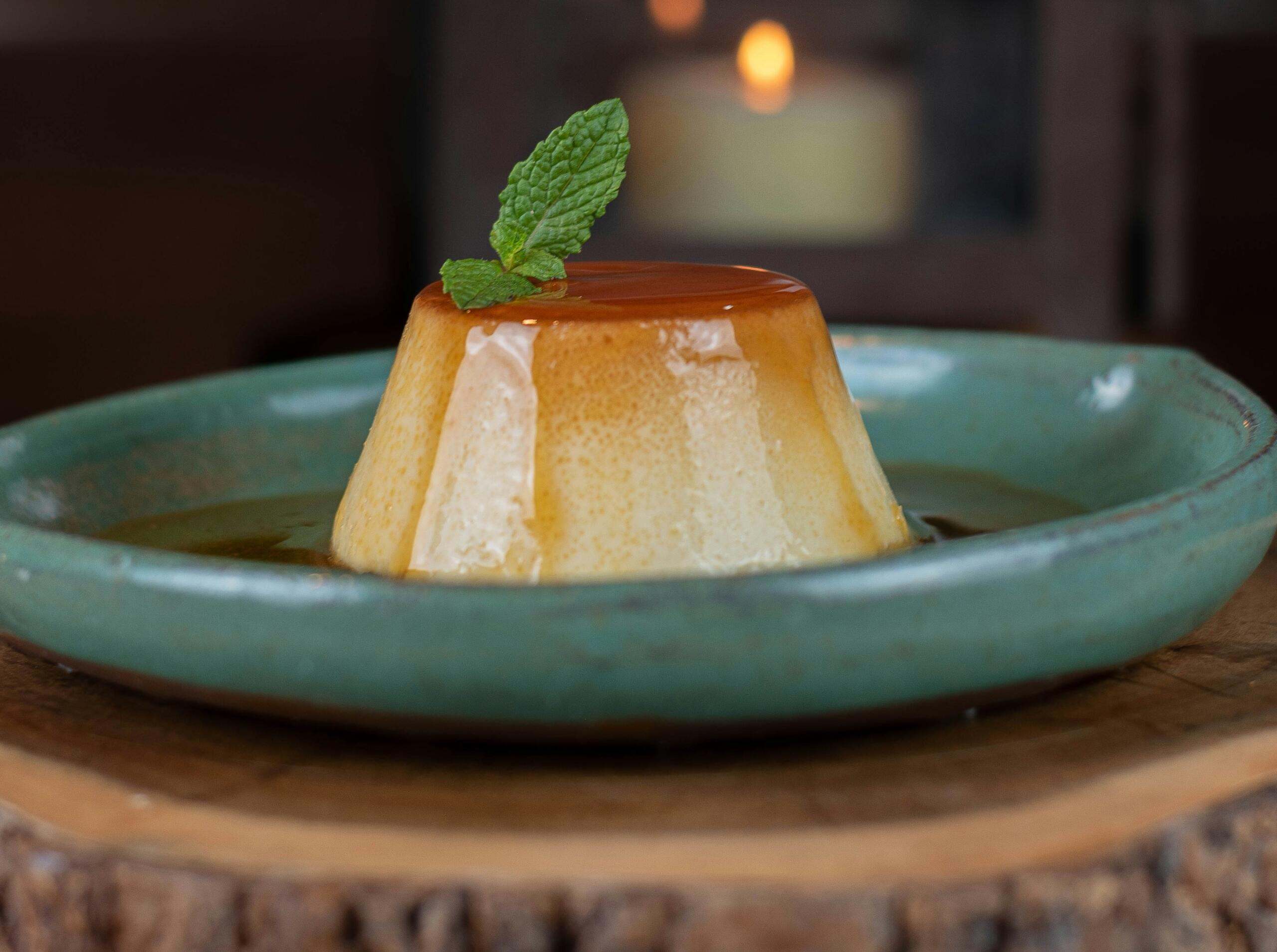 Un cremoso flan de caramelo descansa elegantemente sobre un plato verde, adornado con una hoja de menta y rodeado de un suave y cálido resplandor.