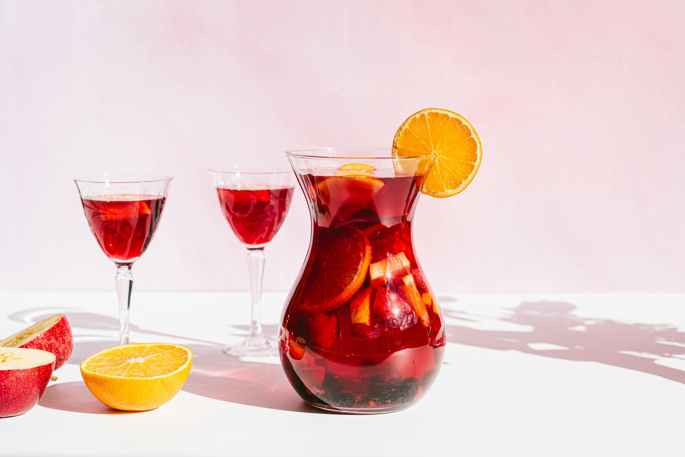 Jarra sangria una bebida con vino tinto y frutos rojos adornada con rodajas de naranja, acompañada de elegantes copas y fruta fresca sobre una superficie blanca.