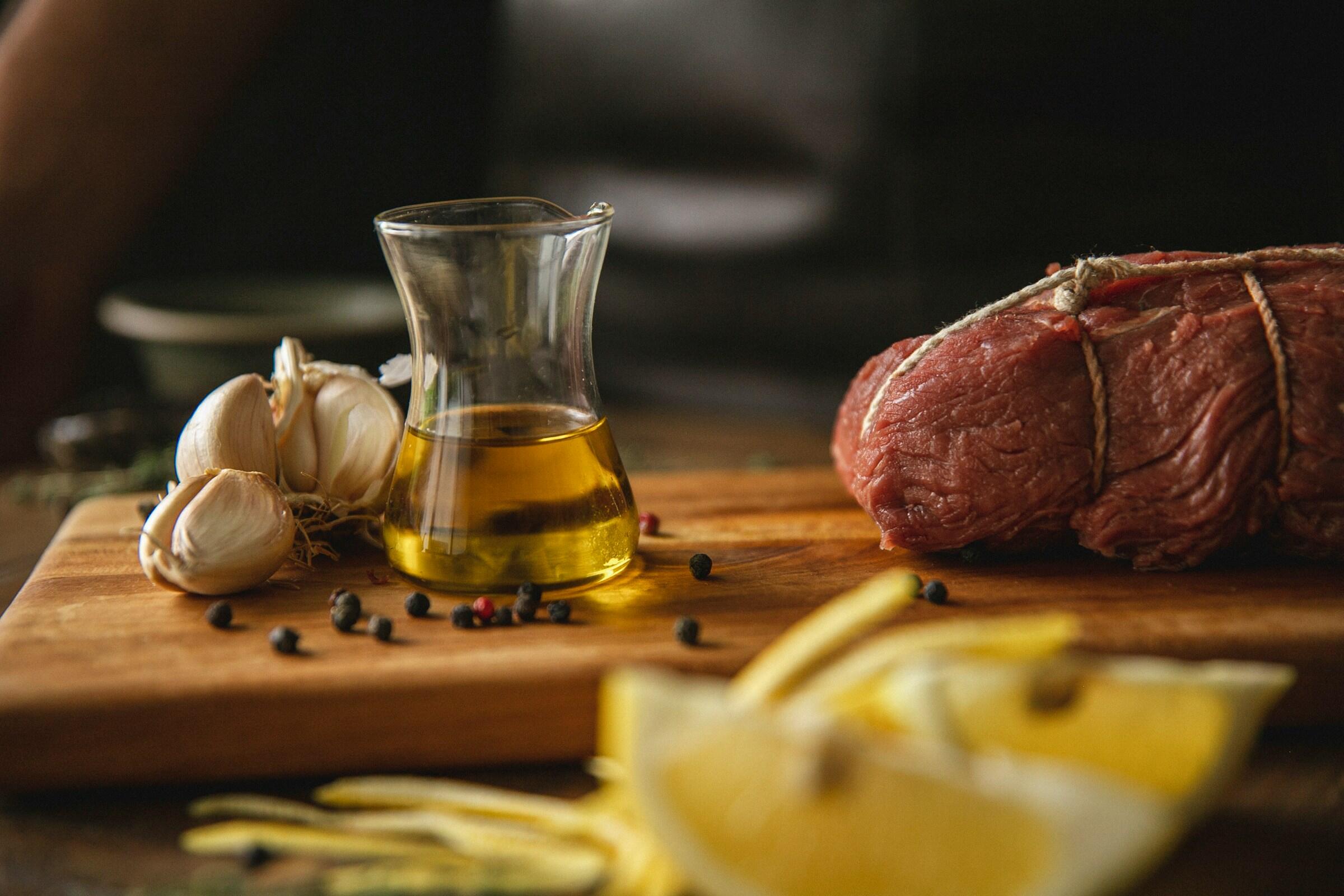 Una tabla de cortar de madera muestra ajo fresco, una botella de vidrio de aceite de oliva, carne cruda atada y rodajas de limón listas para preparar