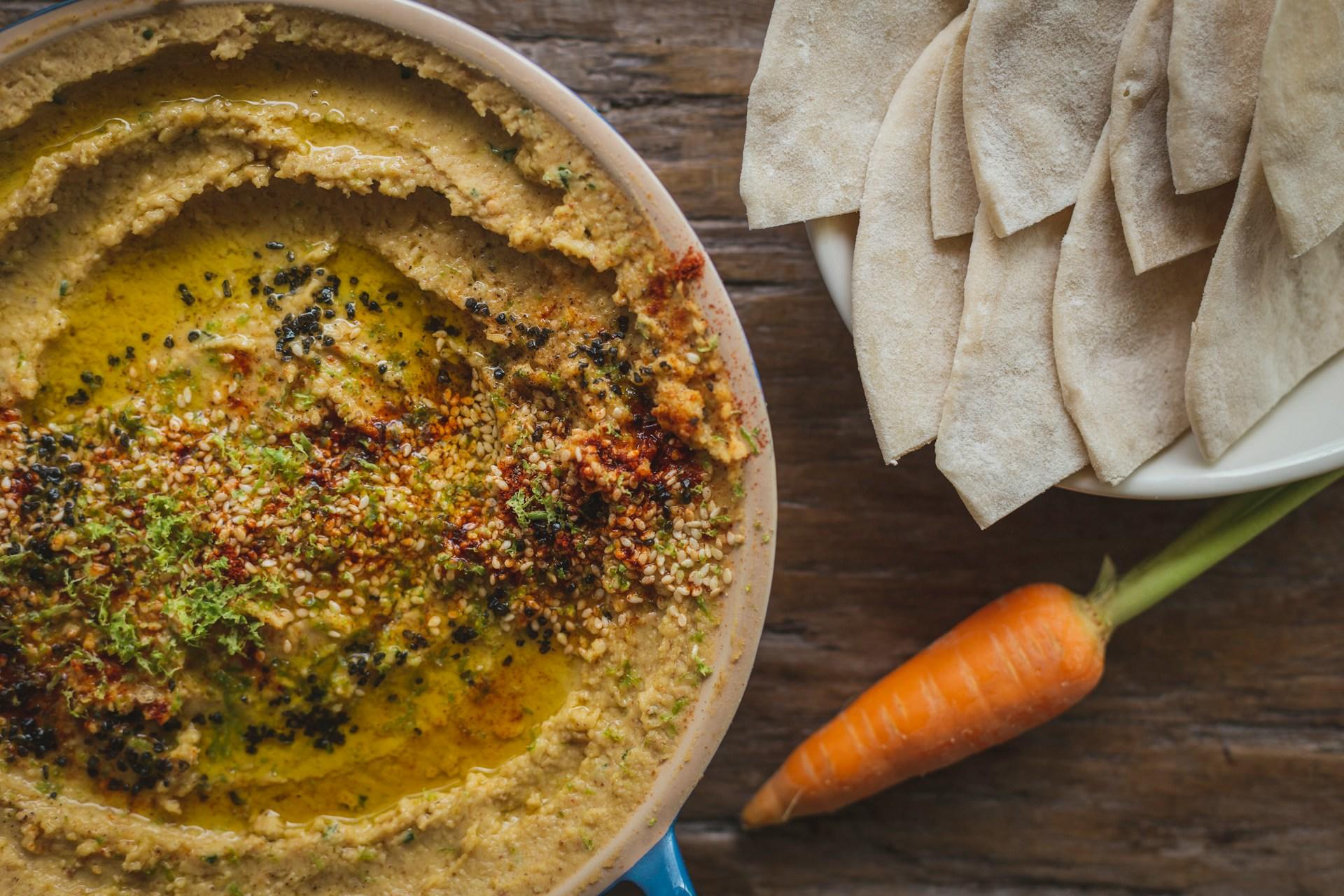 un bowl de humus decorado con semillas y pan pita al costado derecho con una zanahoria
