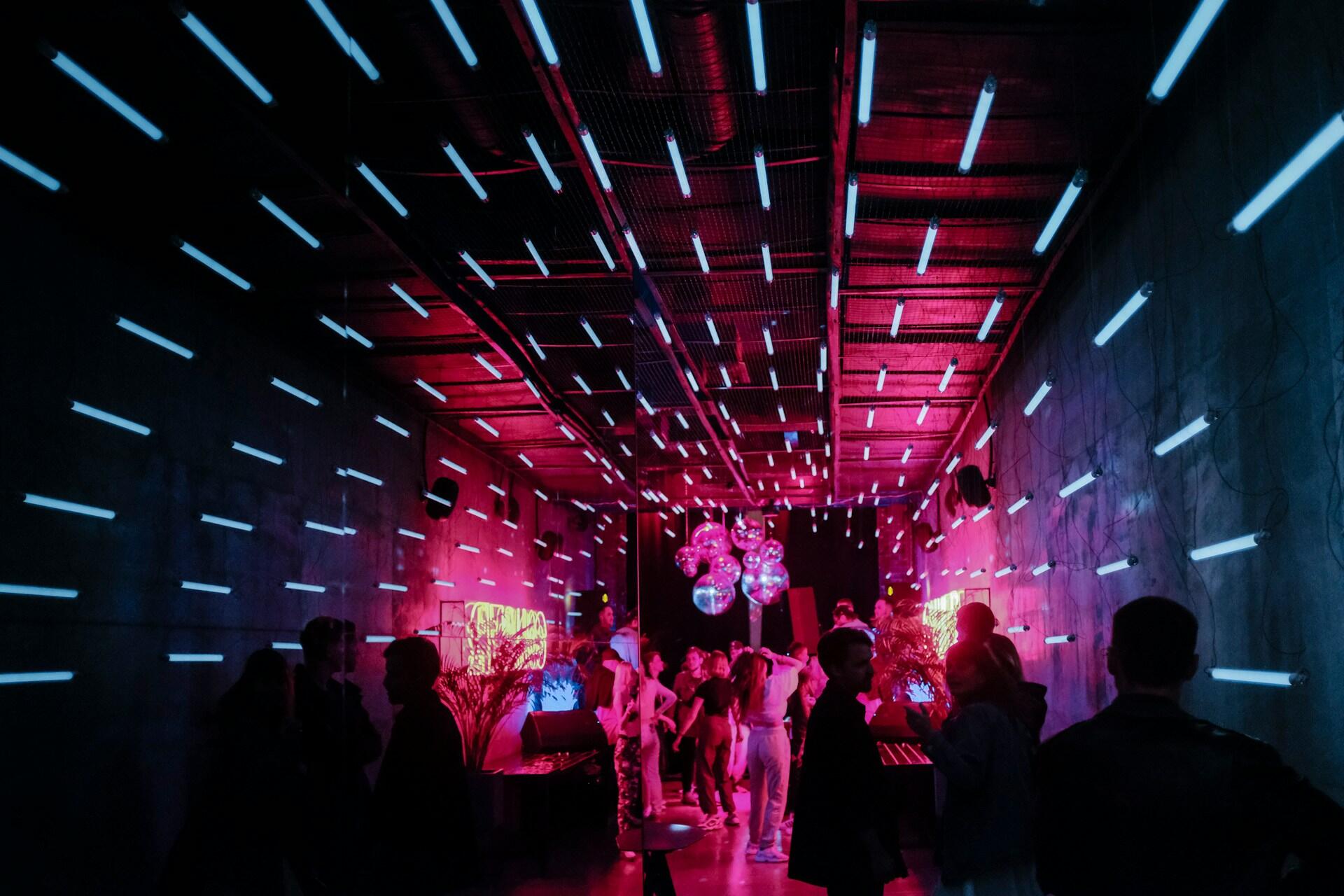 Sala de fiestas con iluminación tenue y luces de neón azules y rosas. La gente se reúne y charla bajo bolas de discoteca colgantes, creando un ambiente enérgico y animado.