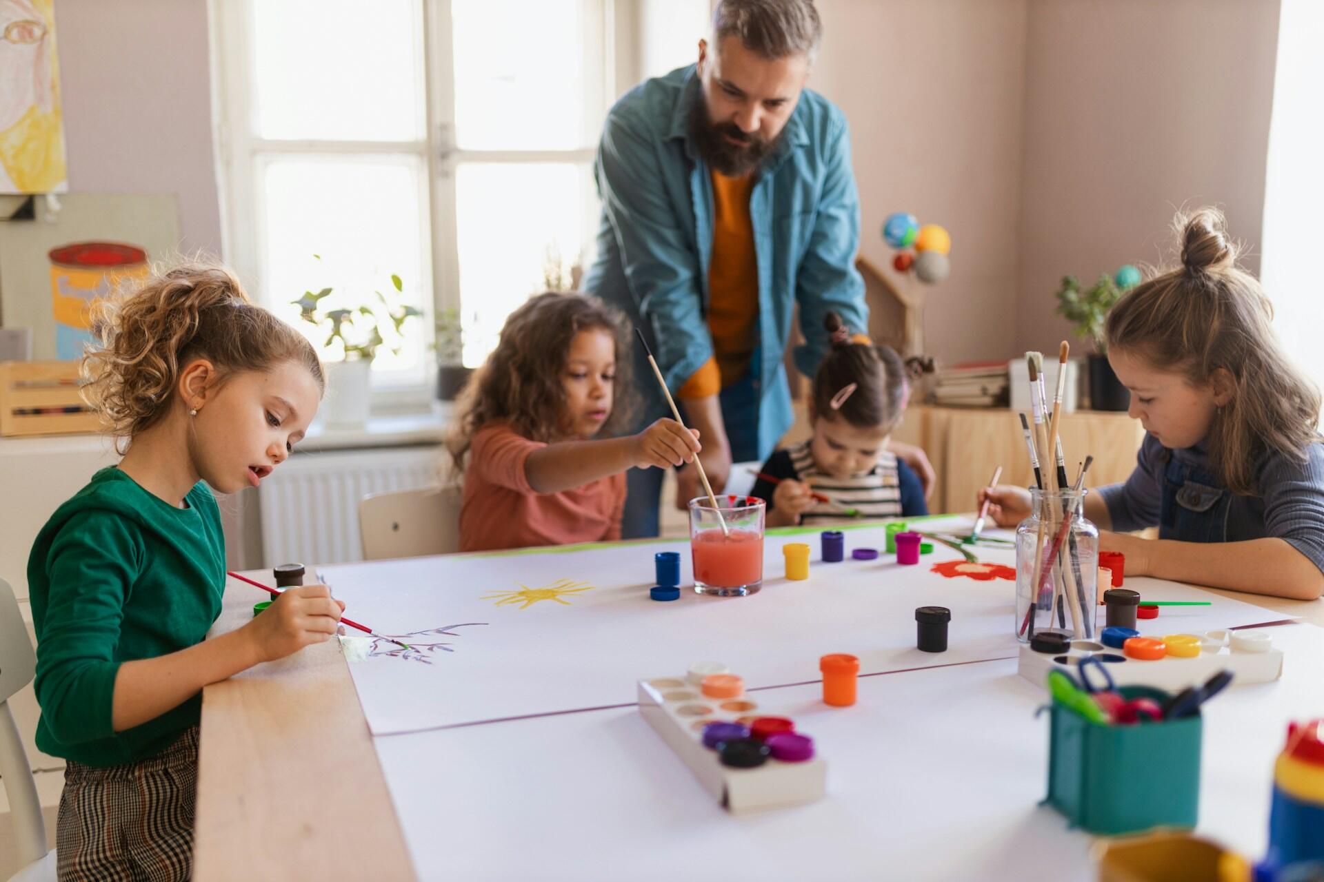 Niños pintando en una mesa con materiales de colores en una habitación muy iluminada.