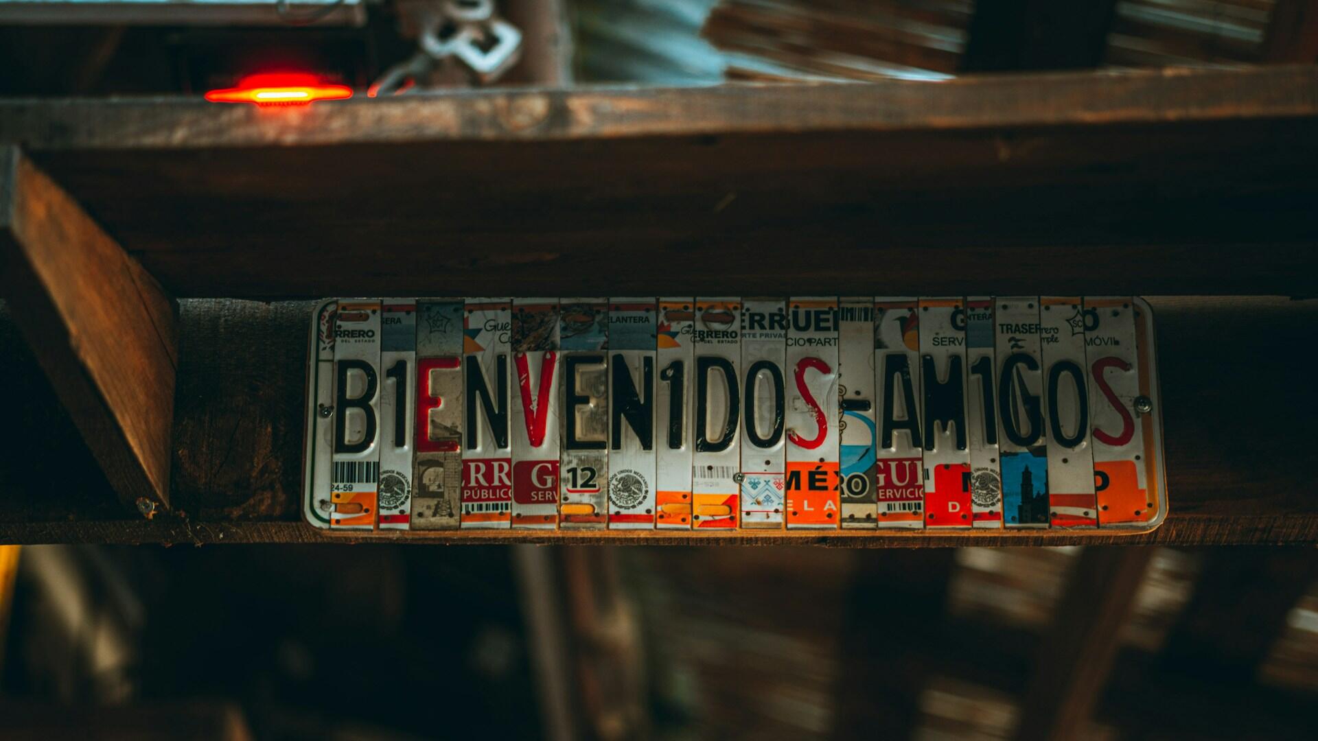 Un letrero creativo, formado por matrículas vintage de colores superpuestas, reza «Bienvenidos, amigos». Está colocado sobre una viga de madera rústica, lo que le da un aire acogedor.