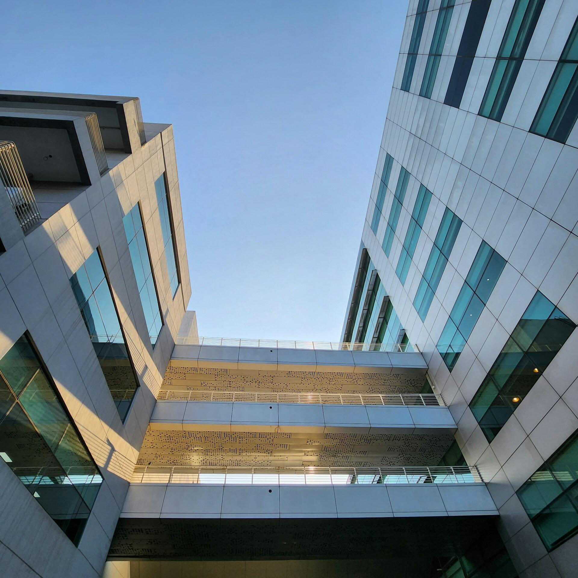 Vista hacia arriba entre modernas torres de oficinas de cristal y acero, unidas por pasarelas horizontales, que enmarcan un cielo azul y despejado; las líneas geométricas transmiten una sensación de elegancia e imponencia.
