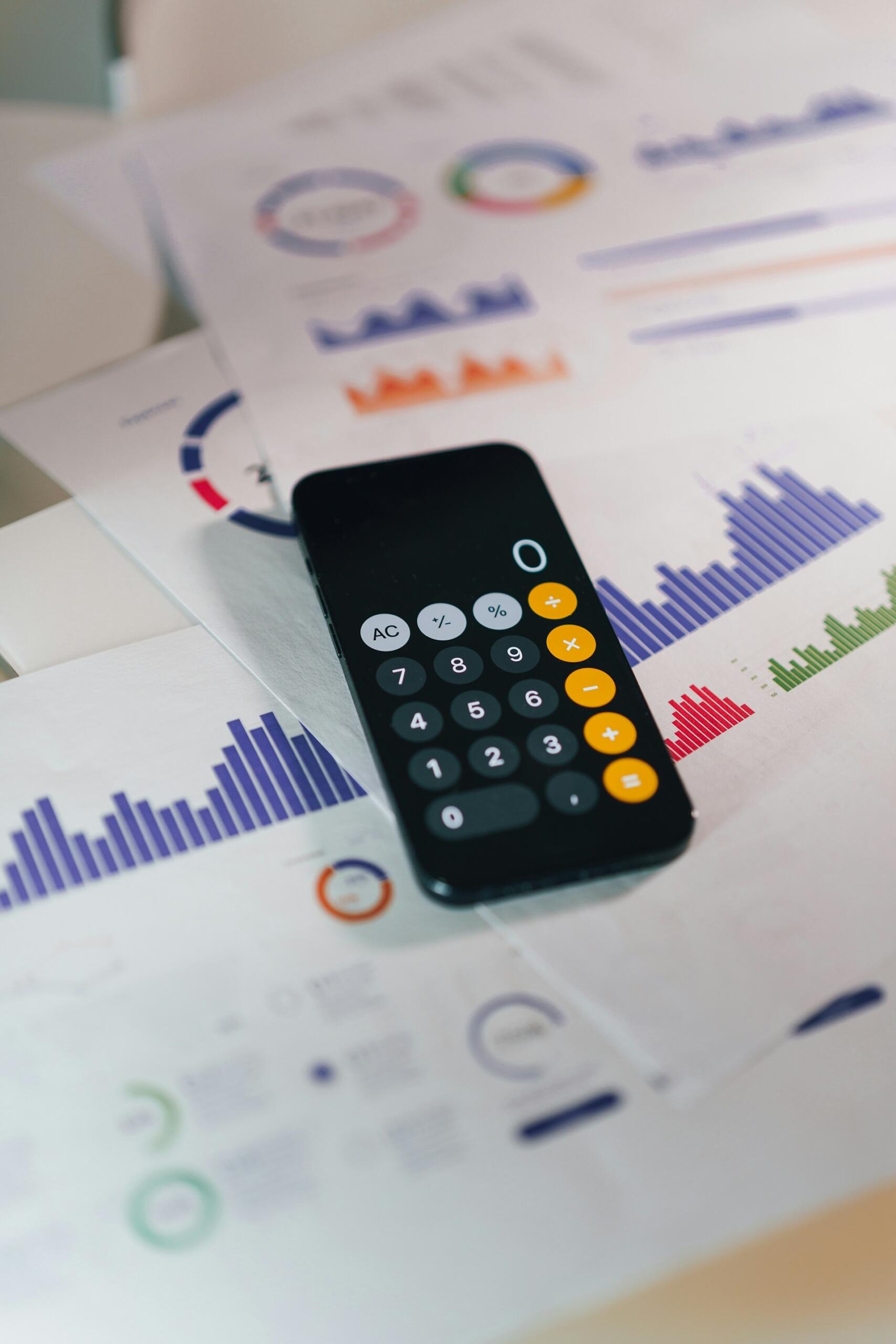 Un smartphone negro en el que se ve una calculadora descansa sobre unos informes financieros con tablas y gráficos de colores, lo que sugiere la elaboración de presupuestos, el análisis y la planificación empresarial.