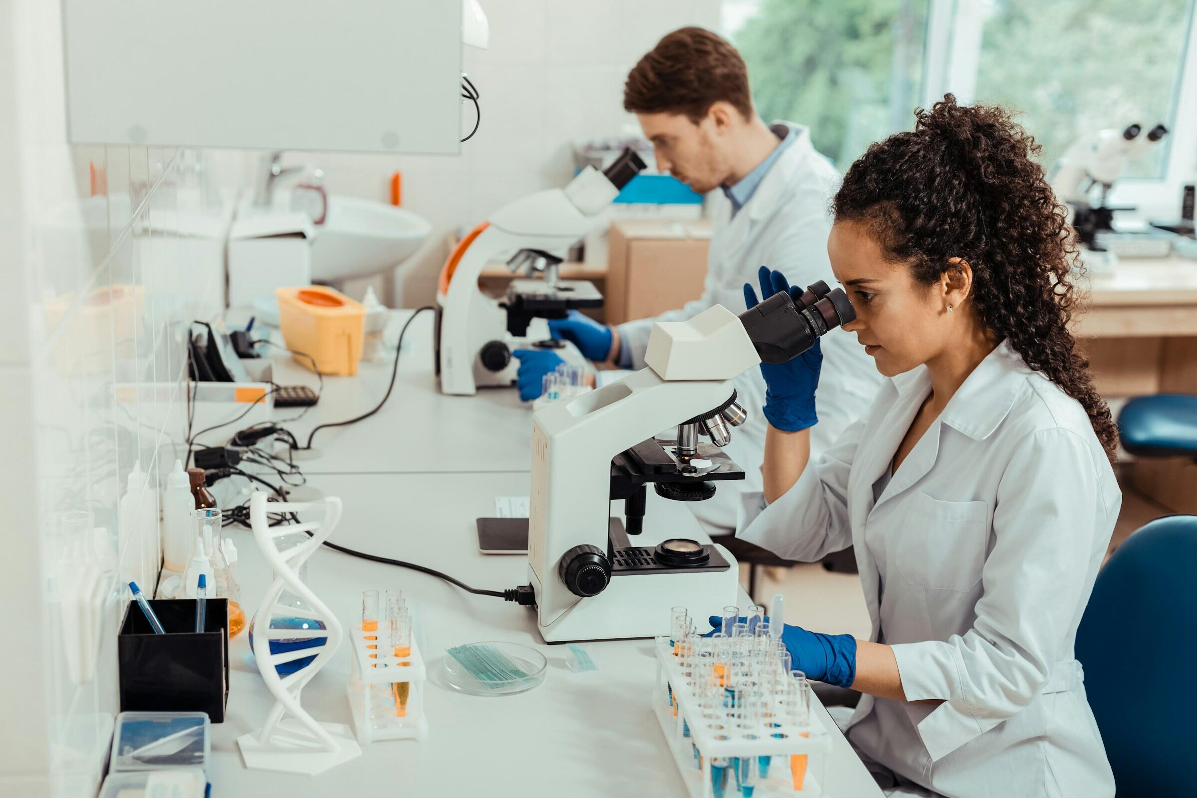 Dos científicos con bata de laboratorio que utilizan microscopios entre aparatos de laboratorio y tubos de ensayo en un luminoso laboratorio.