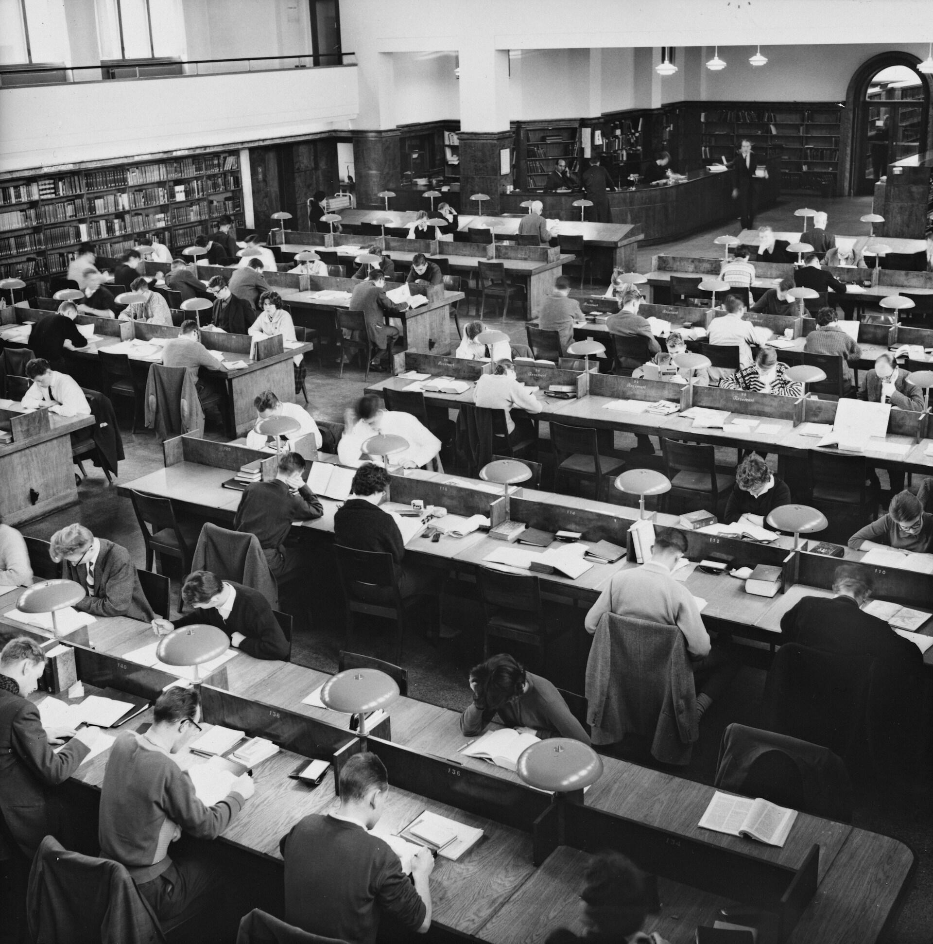 Fotografía en blanco y negro de una sala de lectura abarrotada de una biblioteca, en la que se ve a estudiantes estudiando en largos pupitres bajo lámparas, en un ambiente tranquilo y propicio para el estudio.