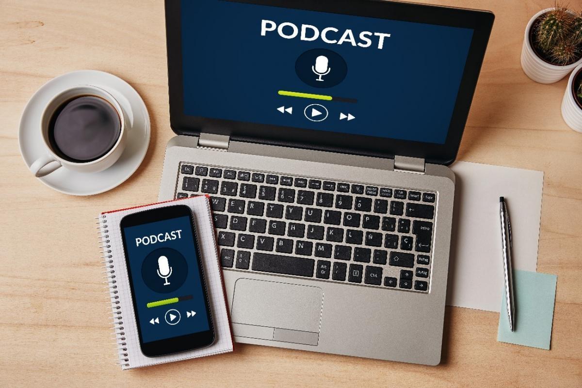 vista cenital de laptop y celular reproduciendo un podcast a la vez junto a unas hojas, lapicero y una taza de cafe