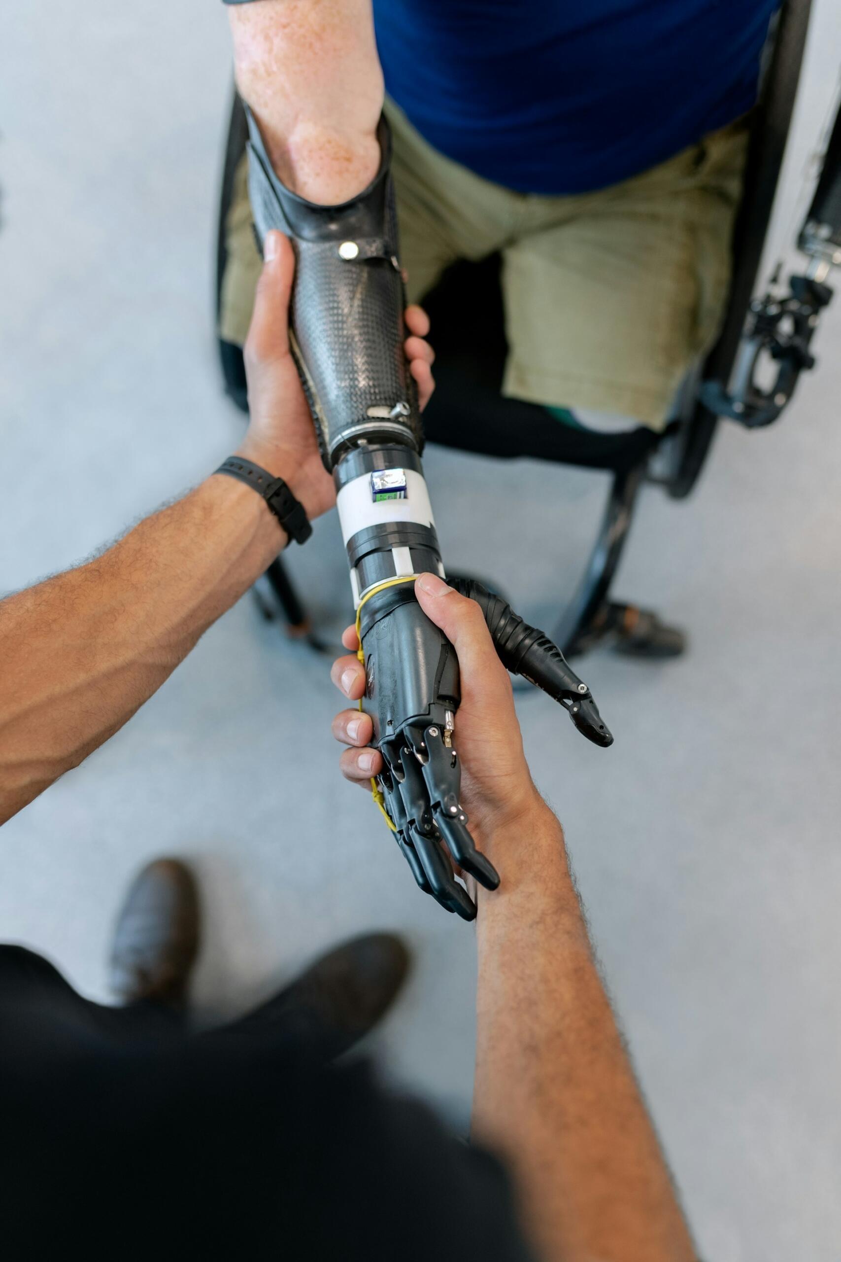 Dos manos conectan un brazo protésico a una persona sentada en una silla de ruedas. La escena transmite colaboración y apoyo, y pone de relieve la avanzada tecnología protésica.