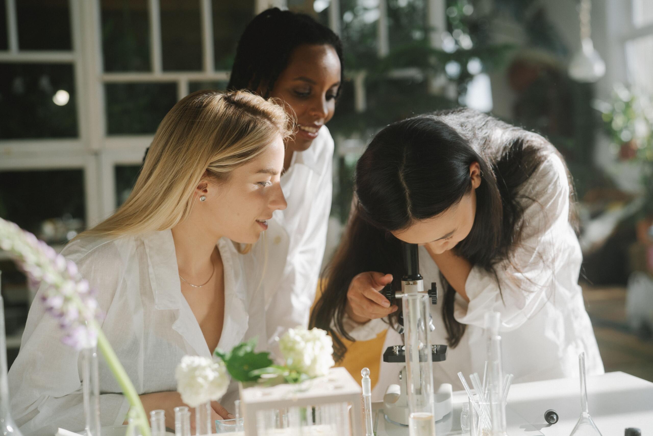 Tres mujeres en un luminoso laboratorio: una mira por el microscopio mientras las demás la observan, rodeadas de material de laboratorio y plantas.